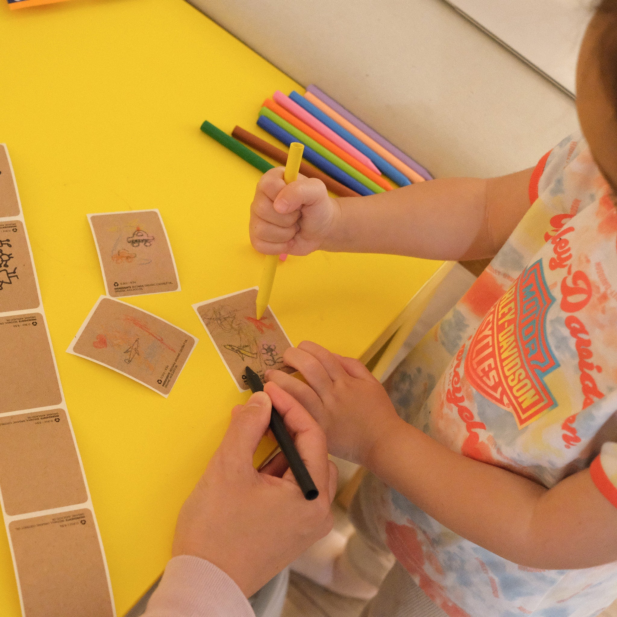 Markers and paper on yellow table, child coloring with yellow marker, Harley-Davidson shirt visible
