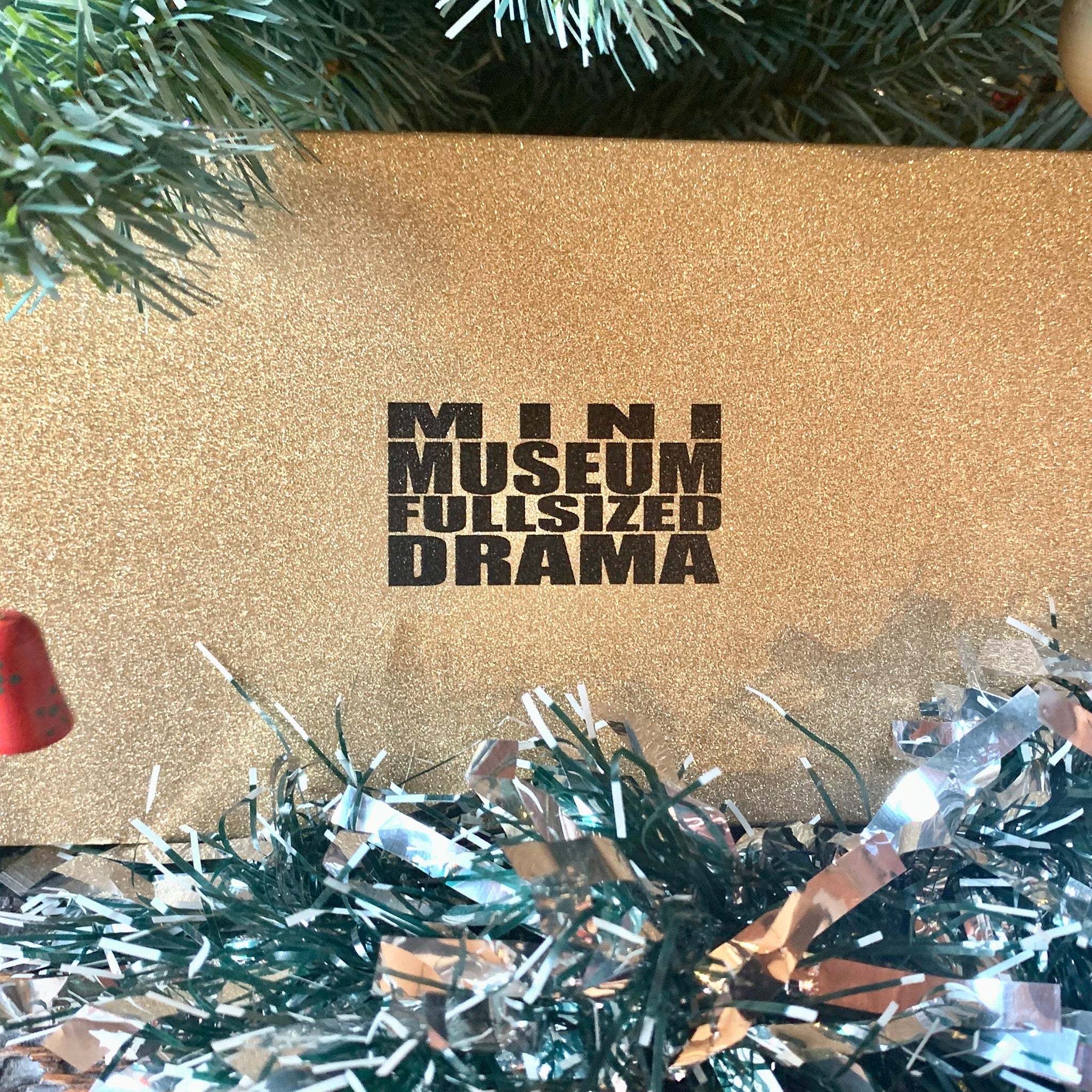 Gold glittery box with black text "MINI MUSEUM FULLSIZED DRAMA" on a Christmas background