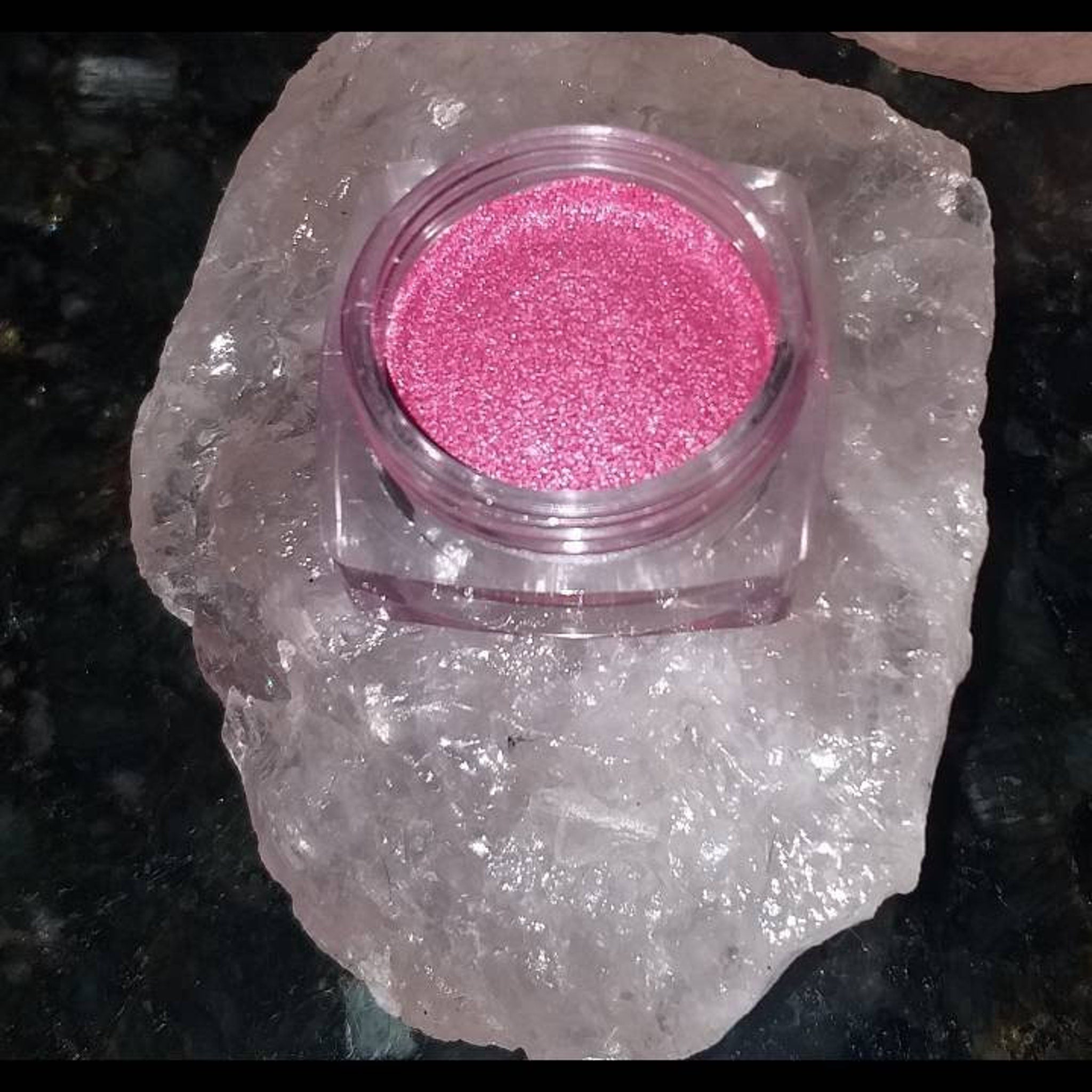Pink glitter in a clear jar on a white crystal stone, close-up shot on a dark surface