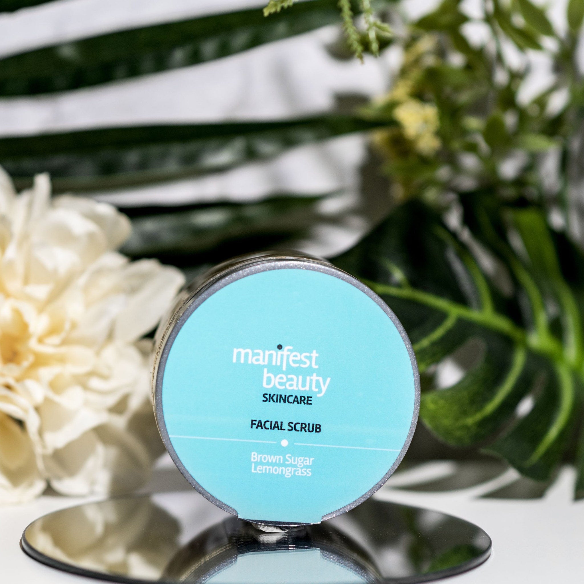 Facial scrub jar with blue label, silver rim, and "Manifest Beauty" text on a mirrored surface