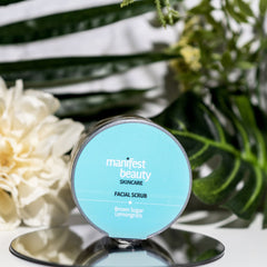 Facial scrub jar with blue label, silver rim, and "Manifest Beauty" text on a mirrored surface