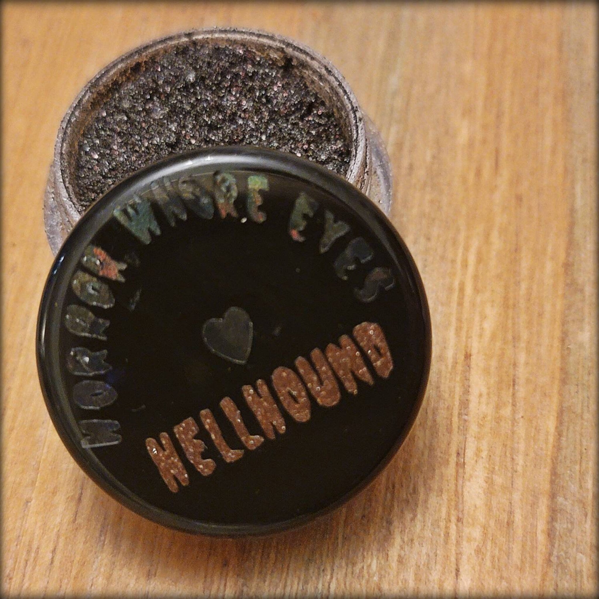 Side view of Hellhound loose eye shadow pigments in a sleek black jar with silver lid, showcasing fine shimmer particles.