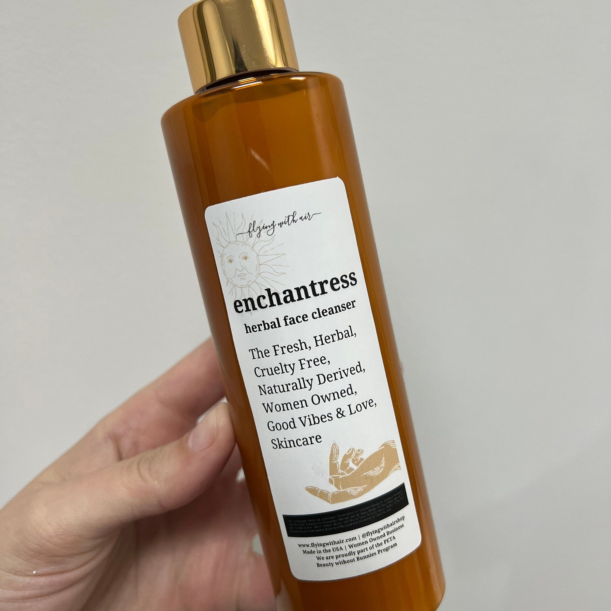 Herbal face cleanser bottle, amber glass, gold cap, "enchantress" label, held in hand
