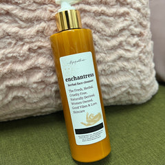 Herbal face cleanser bottle, orange liquid, gold pump, "enchantress" label on green fabric background