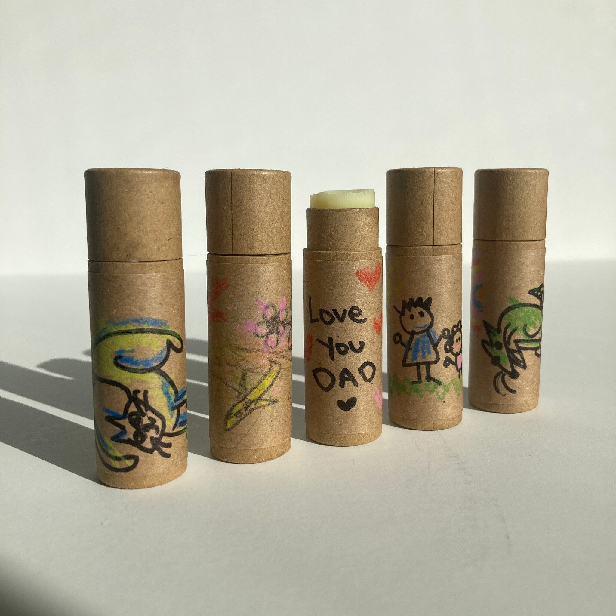 Lip balm tubes with brown paper casing, hand-drawn illustrations, and "Love You DAD" text on one tube