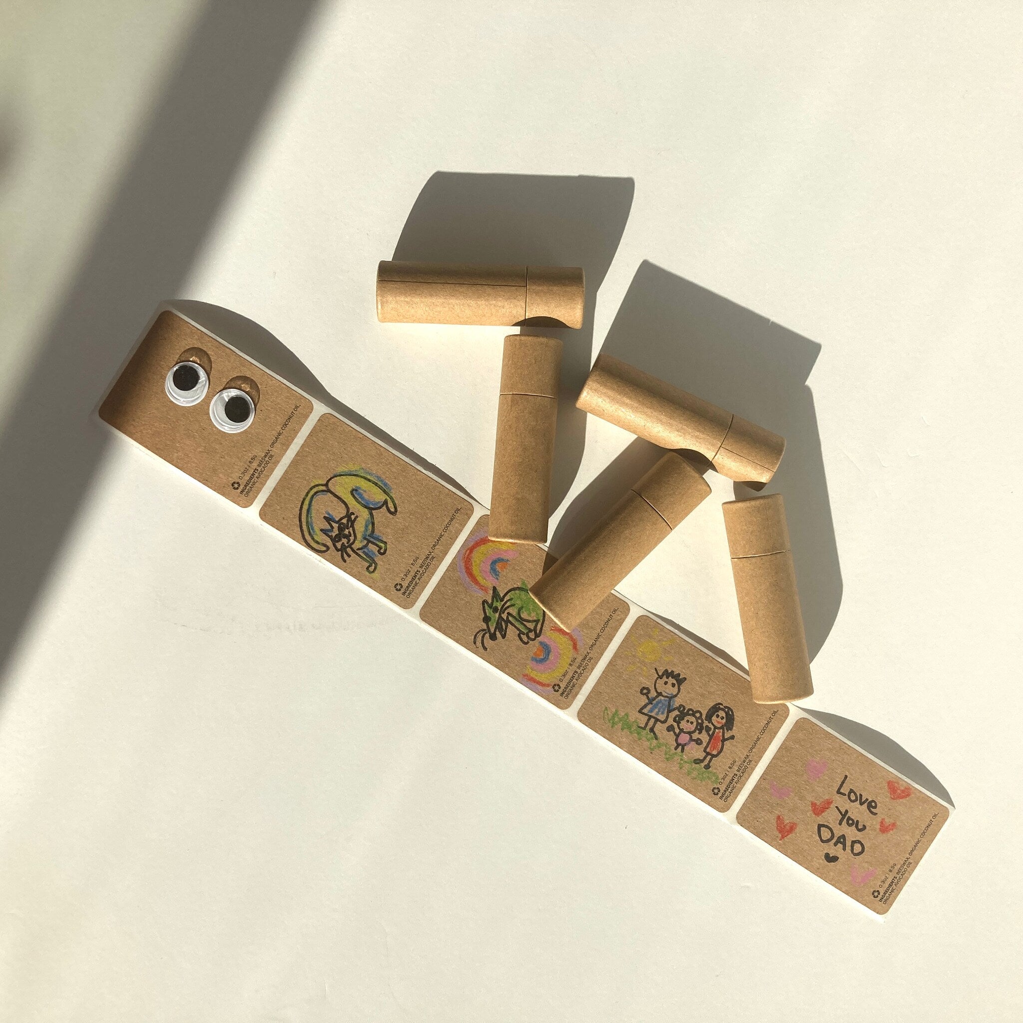 Lip balm tubes with brown paper material and drawings, next to stickers with drawings and text "Love You Dad