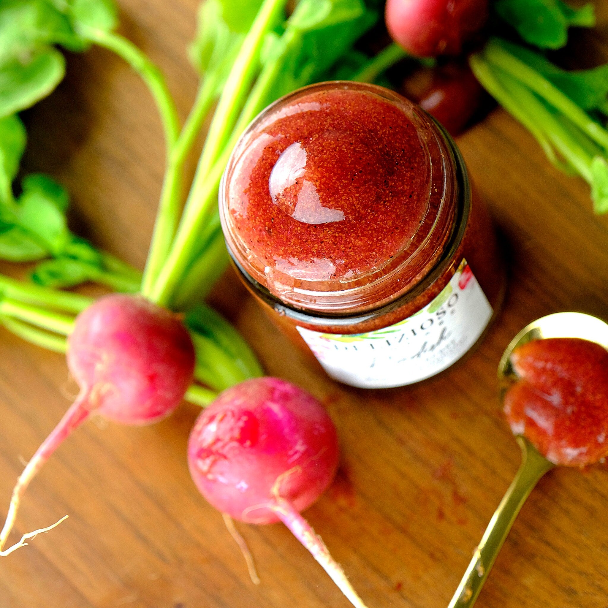 Radish jam in a glass jar, red and speckled, with radishes and green leaves on wood surface