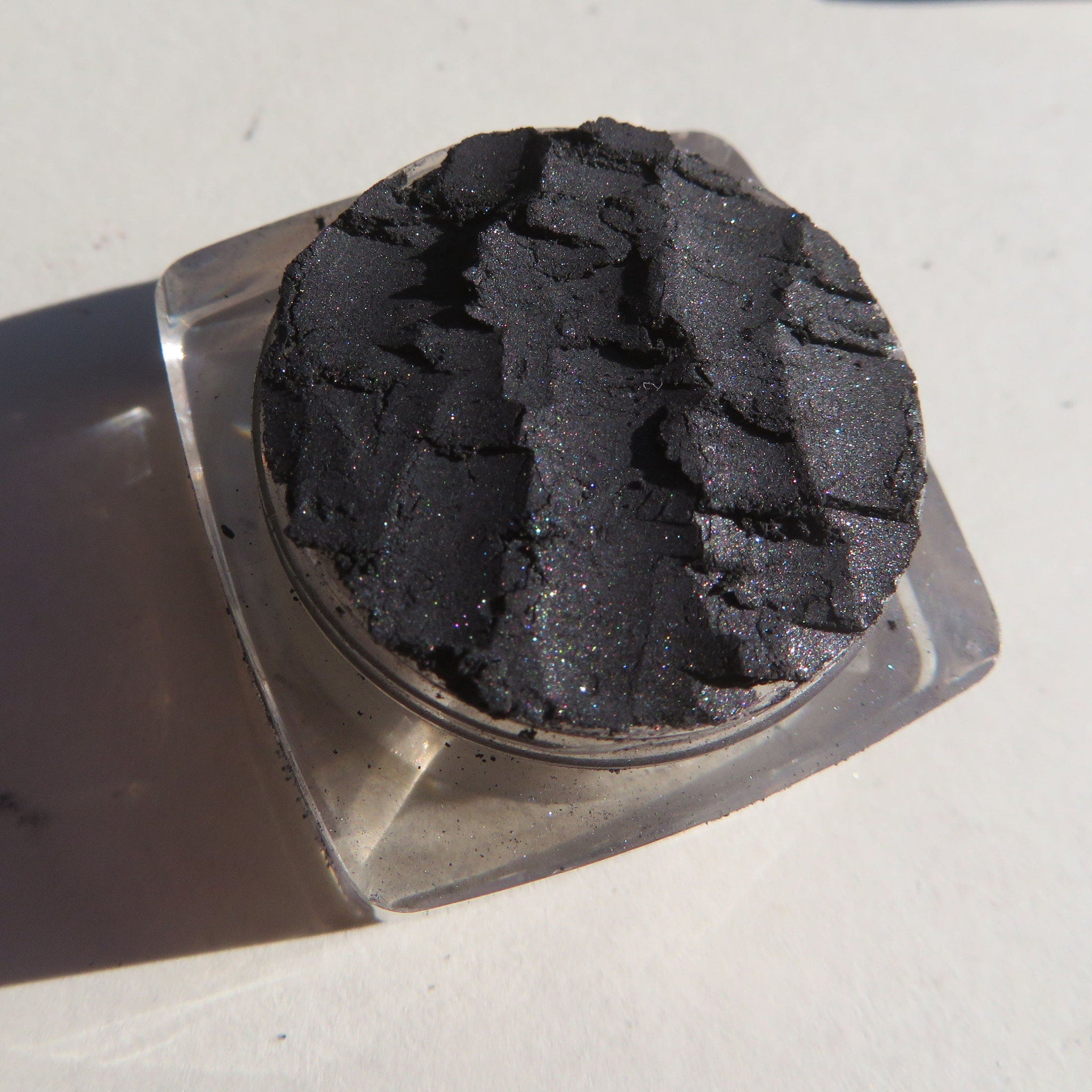 Side view of loose black silver shimmer eyeshadow powder in a clear jar with silver lid, showcasing mineral texture