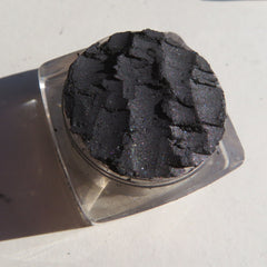 Side view of loose black silver shimmer eyeshadow powder in a clear jar with silver lid, showcasing mineral texture
