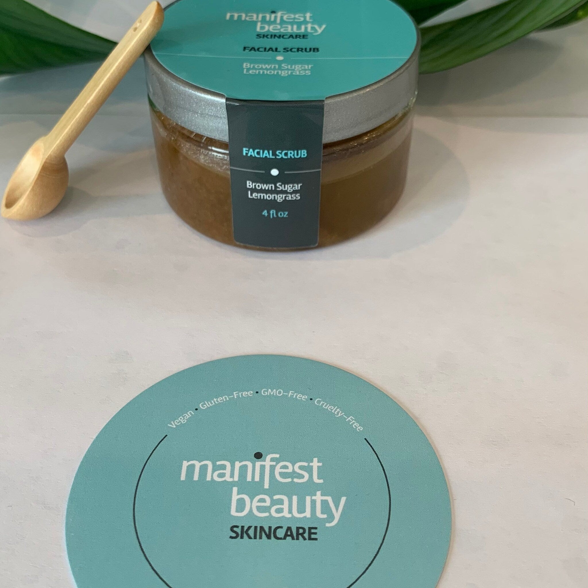 Facial scrub jar with brown contents, silver lid, and wooden spoon near Manifest Beauty label