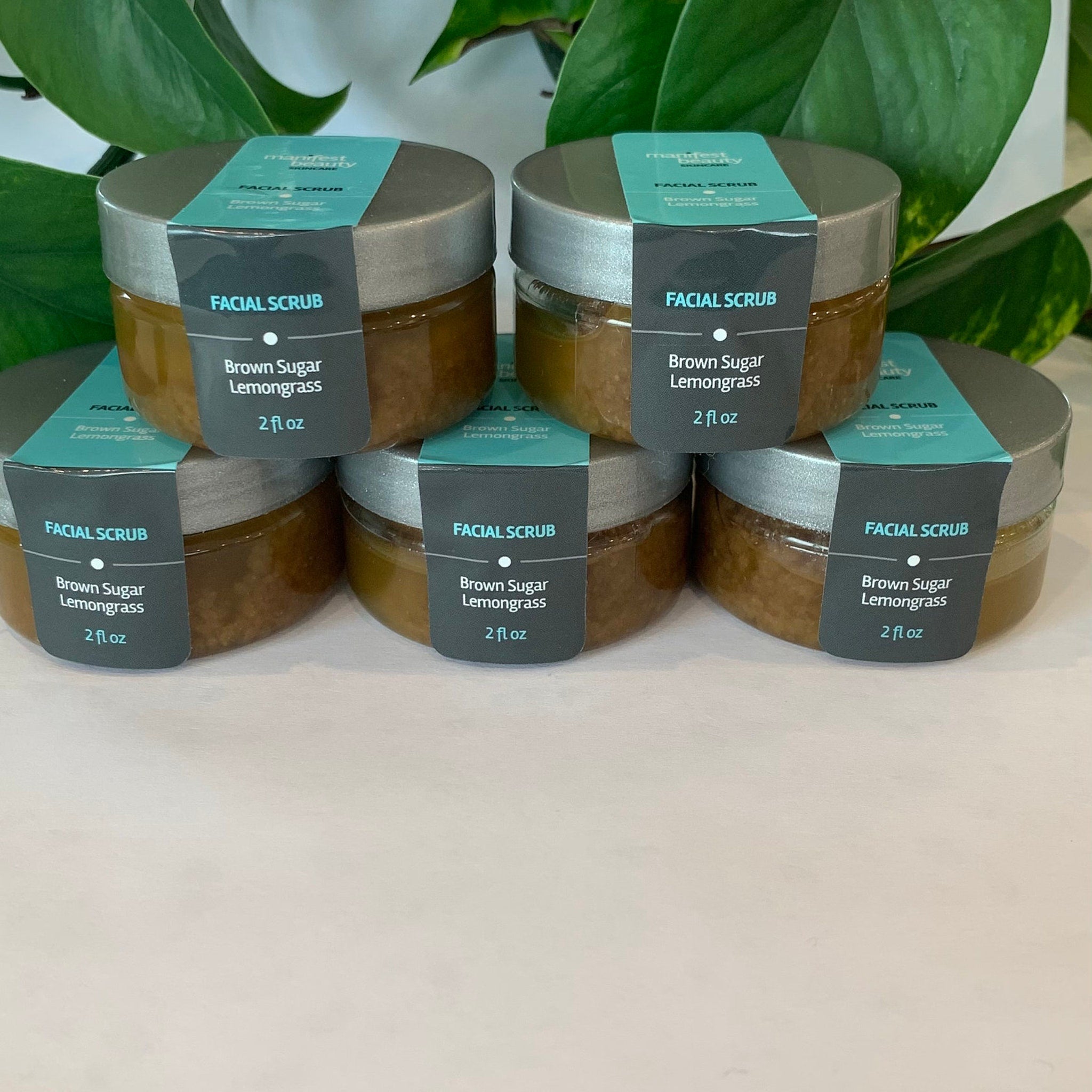 Facial scrub jars with silver lids, brown sugar granules, and Manifest Beauty label on white surface