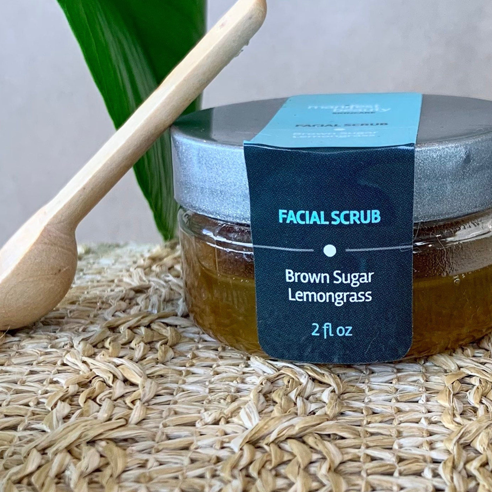 Facial scrub jar with brown liquid, silver lid, and black label, on woven mat with wooden spoon
