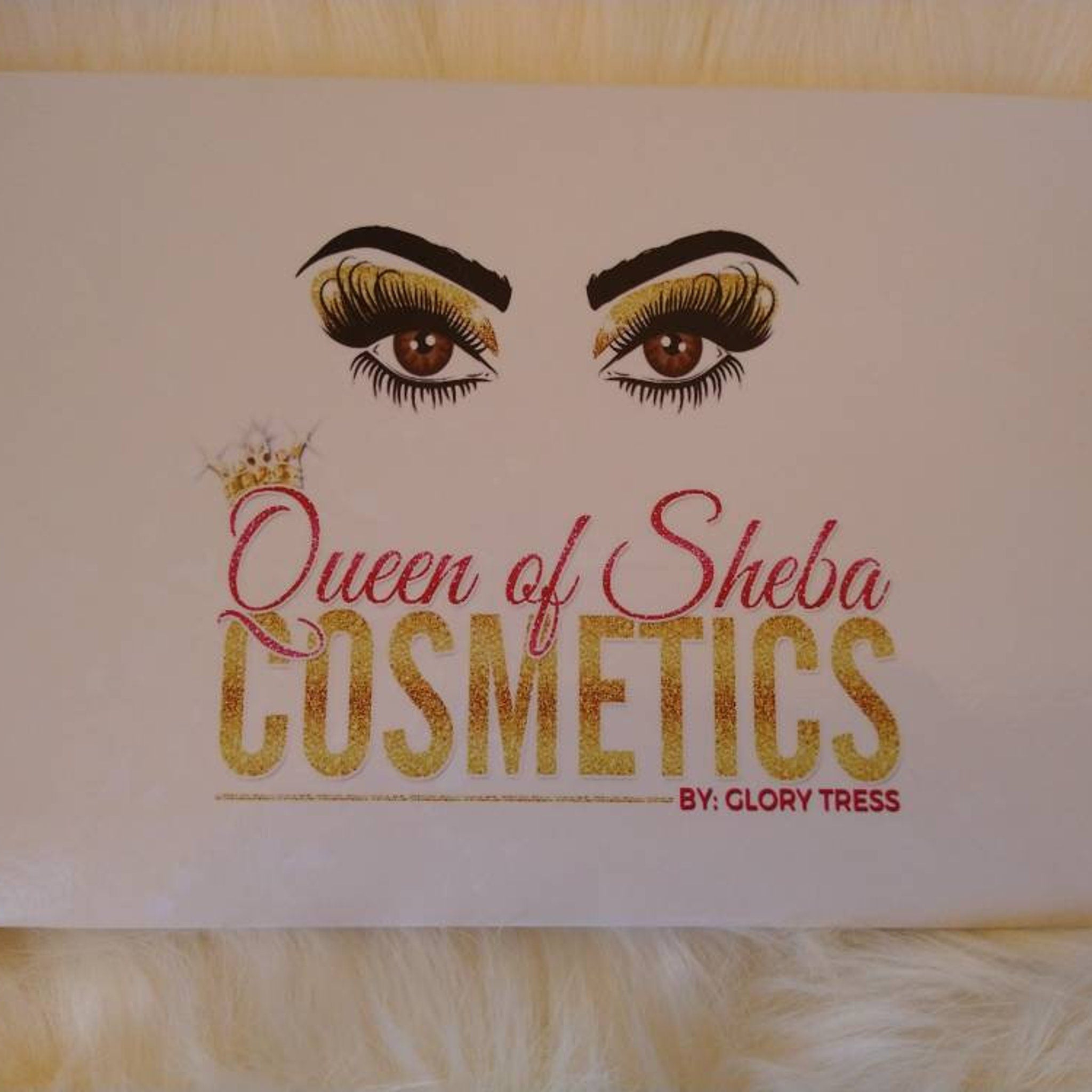 Queen of Sheba Cosmetics logo with brown eyes, gold glitter text, and pink script text on white background
