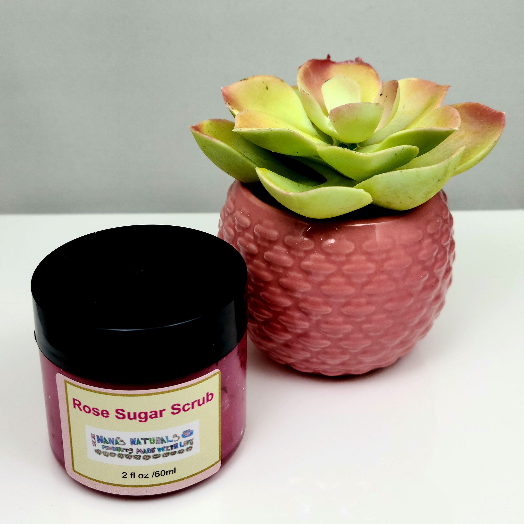 Rose Sugar Scrub jar with pink liquid, black lid, and Nana's Naturals label, next to a pink textured pot with a succulent