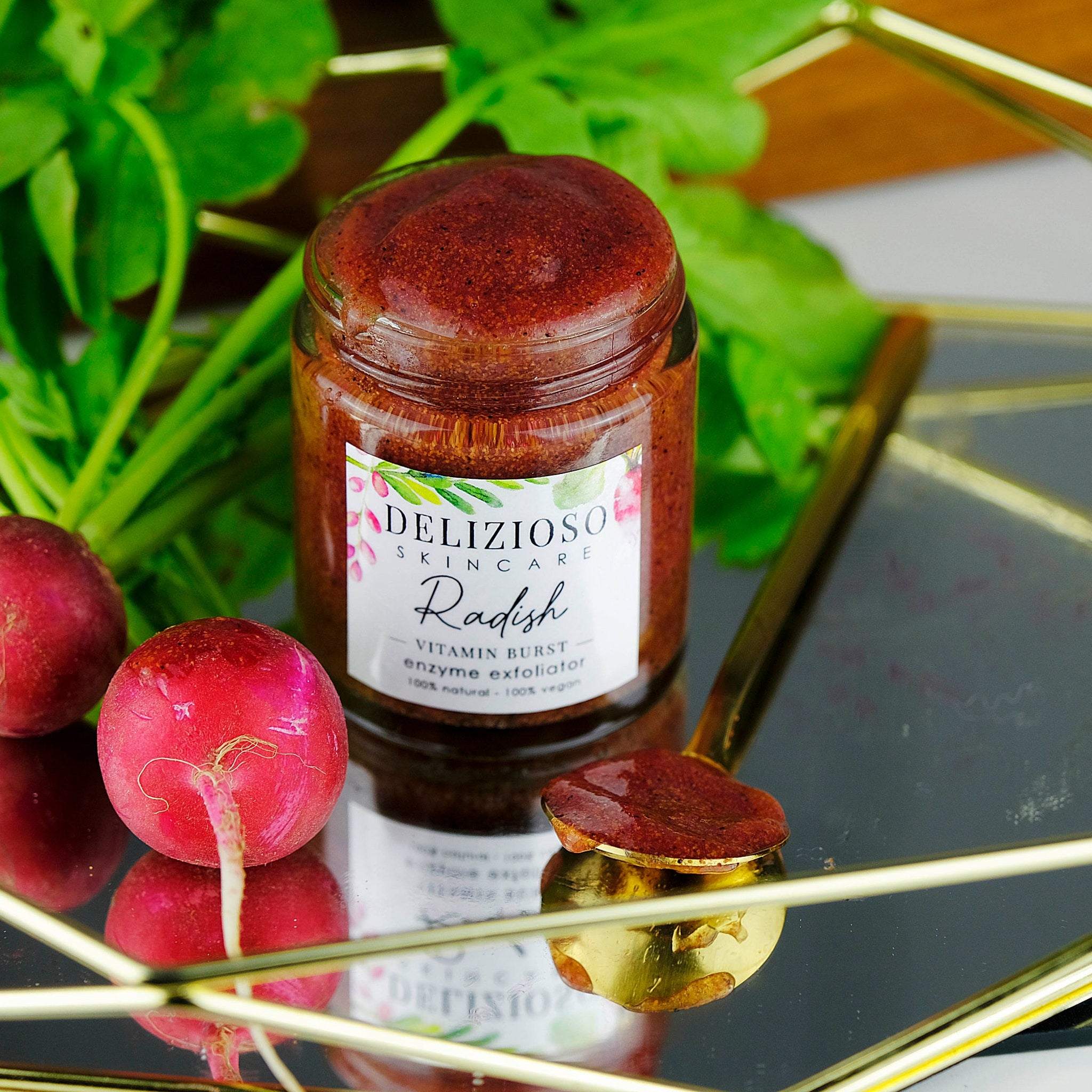 Radish enzyme exfoliator in a glass jar with a brown paste and gold spoon, Delizioso Skincare label visible
