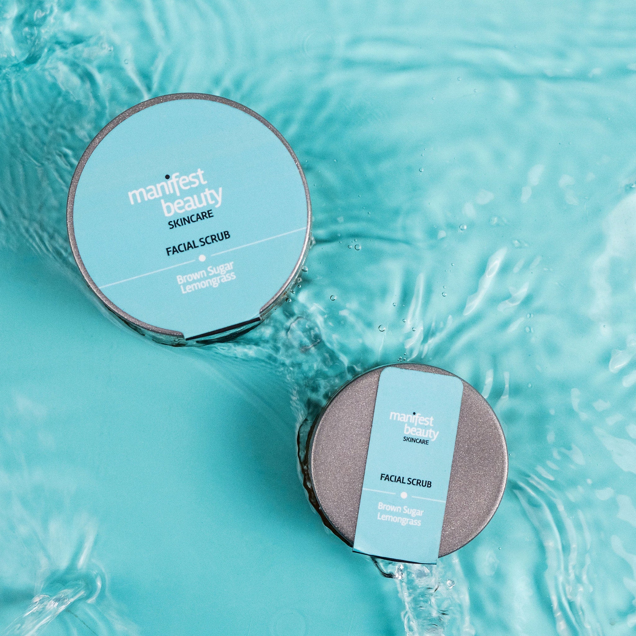 Facial scrub in silver and blue containers with Manifest Beauty branding, floating in water