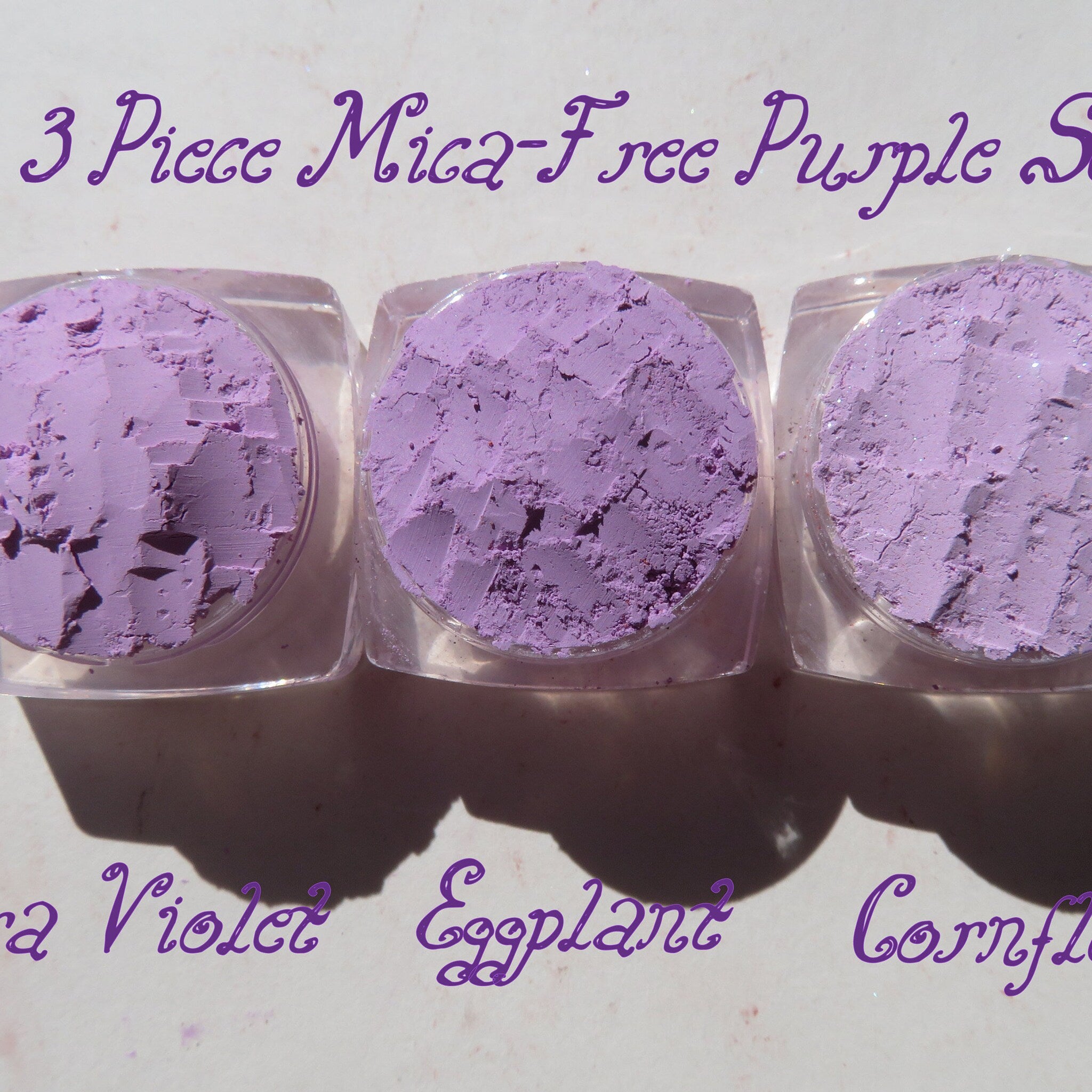 Three loose purple mica eyeshadow pigments in small glass jars with black lids, arranged on a marble surface.