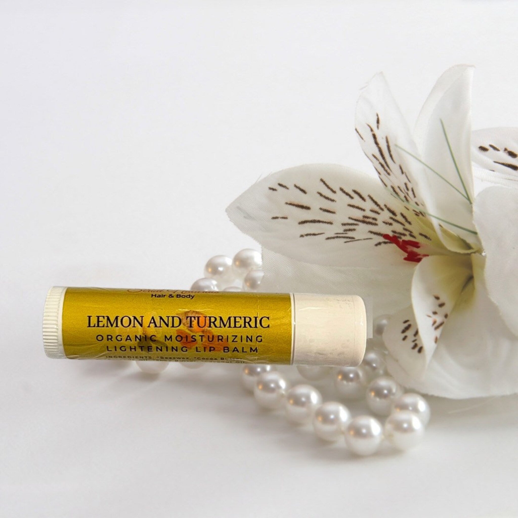 Lip balm tube with yellow label, white flower, and pearl necklace on white surface