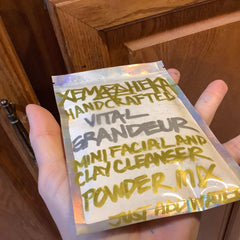 Facial cleanser powder packet, clear foil, white powder, held by hand with text "Xemaheka