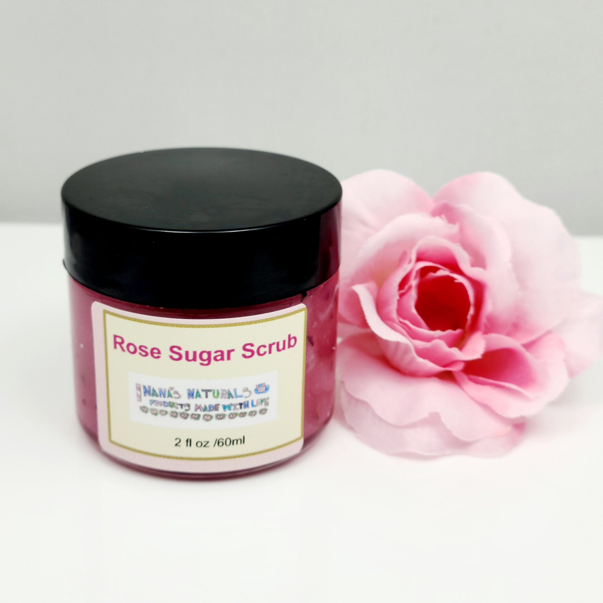 Rose sugar scrub jar with black lid, pink contents, and Nana's Naturals label, next to pink flower