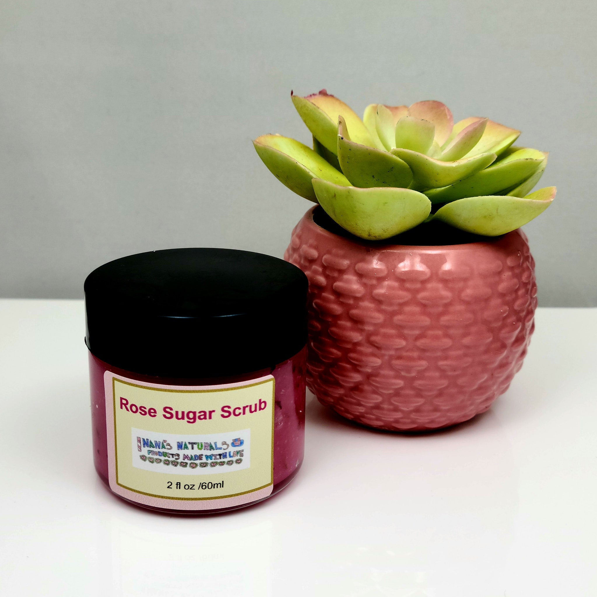 Rose sugar scrub jar with black lid and pink label, next to a pink textured pot with a green plant