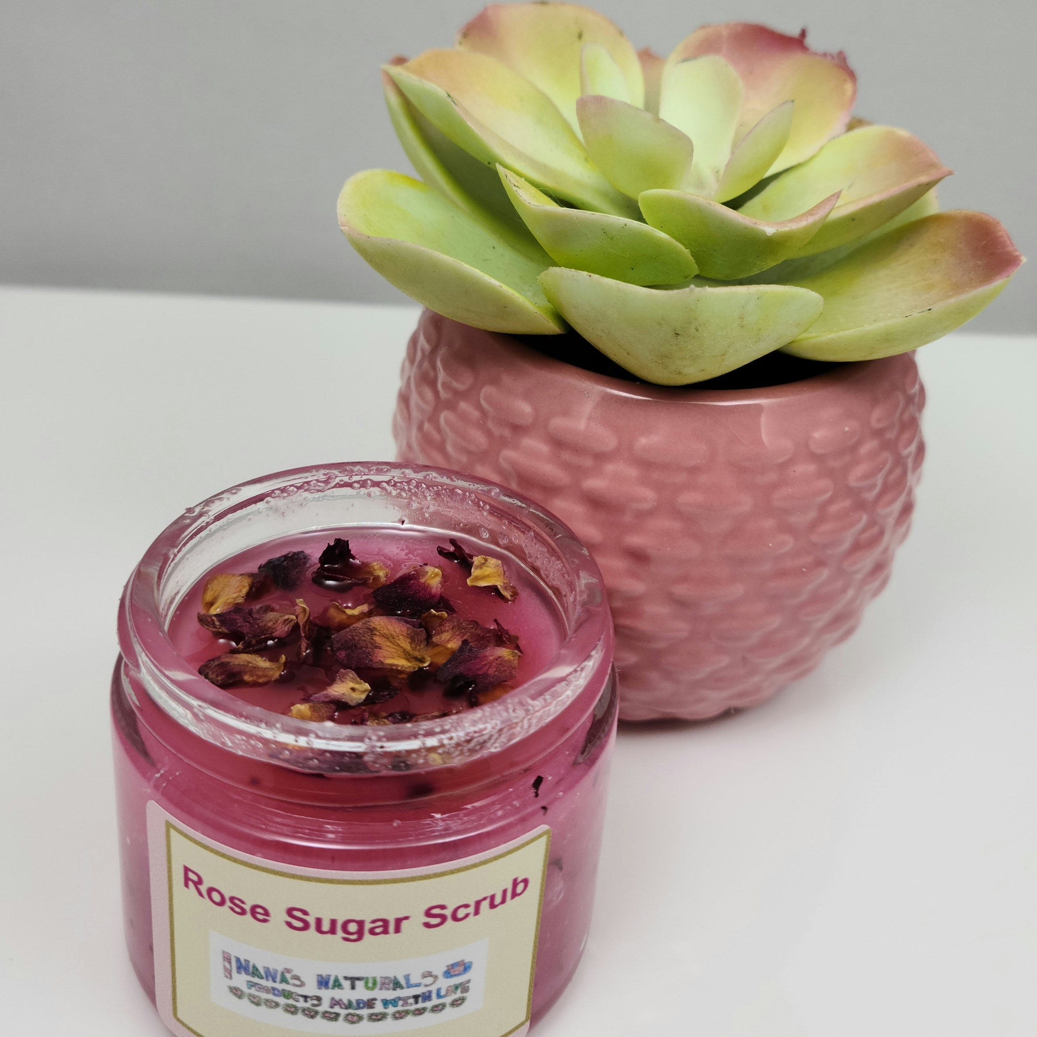 Rose sugar scrub in a clear glass jar with pink liquid and dried rose petals, Nana's Naturals label