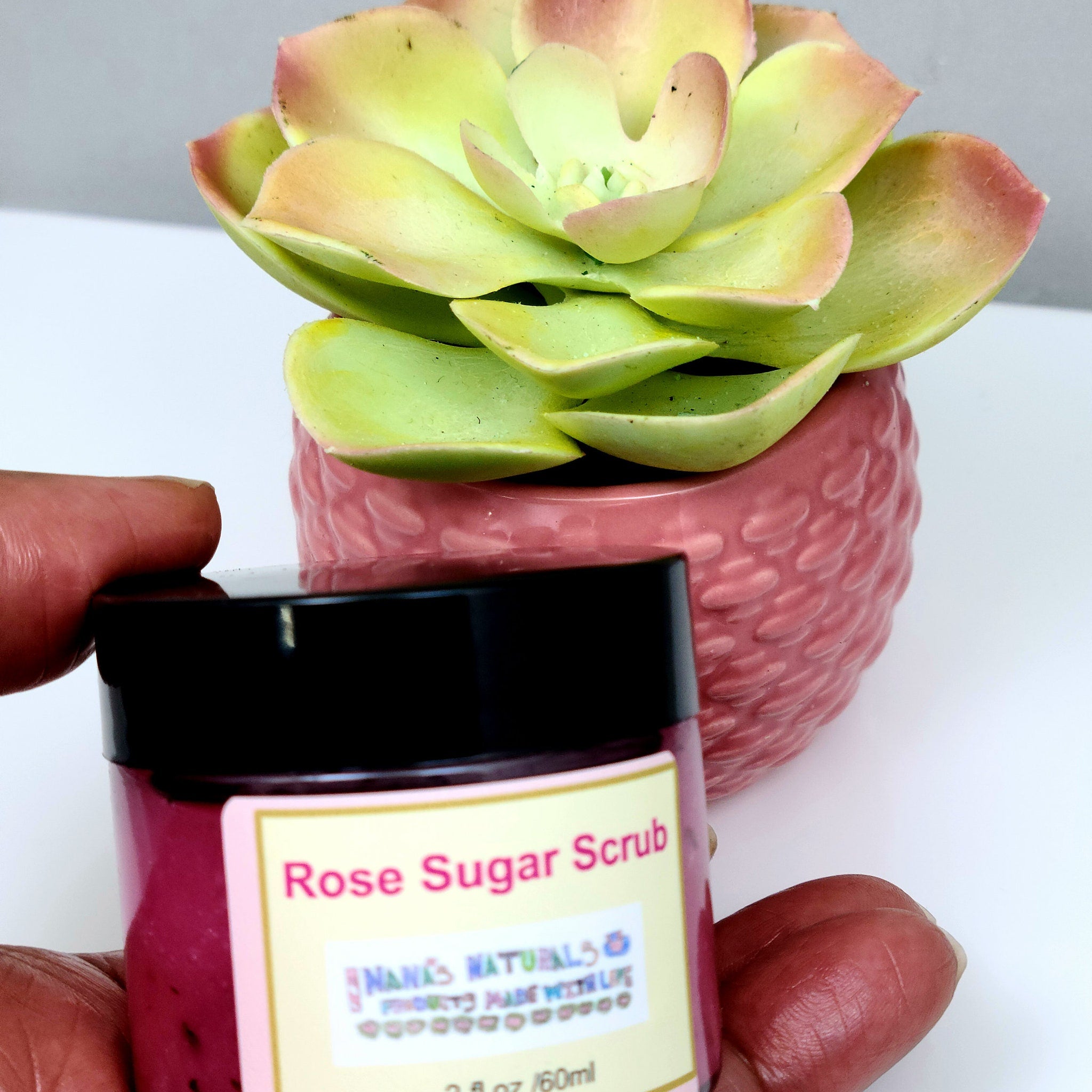 Rose Sugar Scrub jar with pink liquid, black lid, and Nana's Naturals label, held by hand near a succulent plant