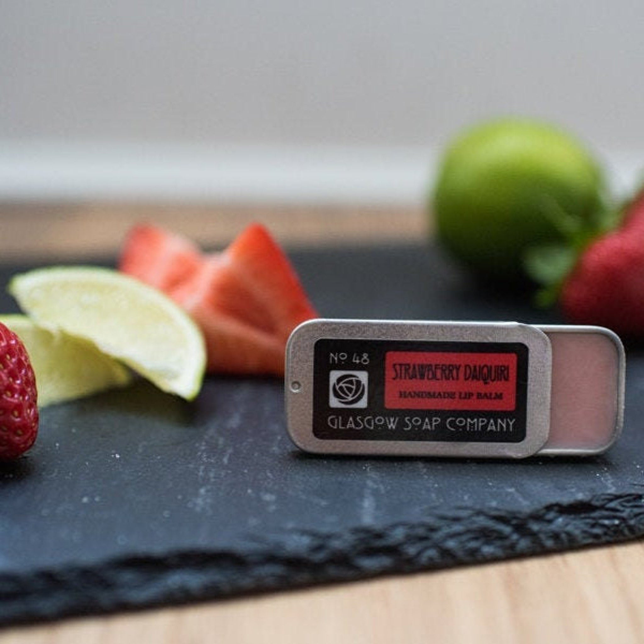 Lip balm in silver tin with pink balm, Glasgow Soap Company branding, and fruit garnish on slate