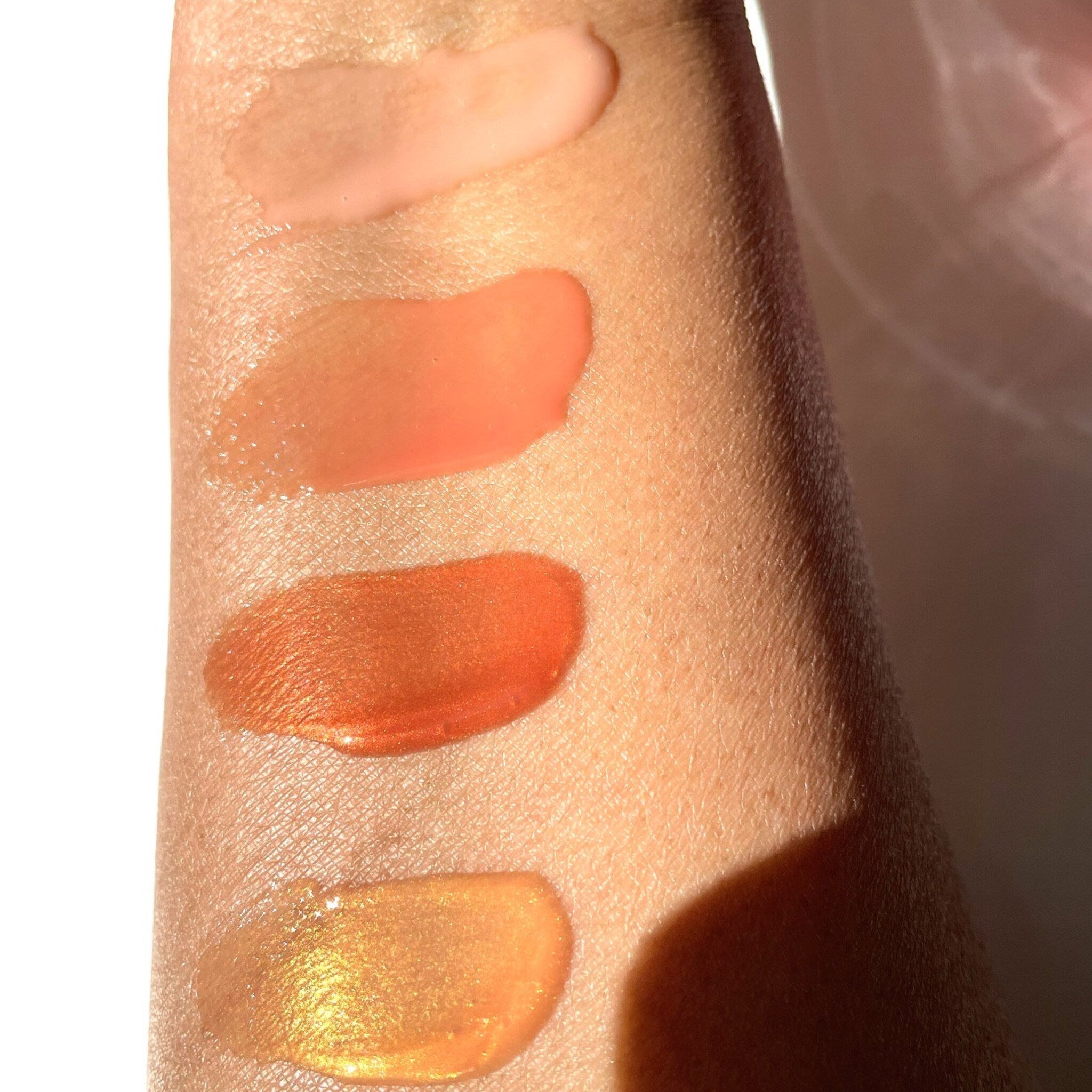 Lip gloss swatches in various shades of pink, orange, and gold on skin, Brownsugarglamcollection