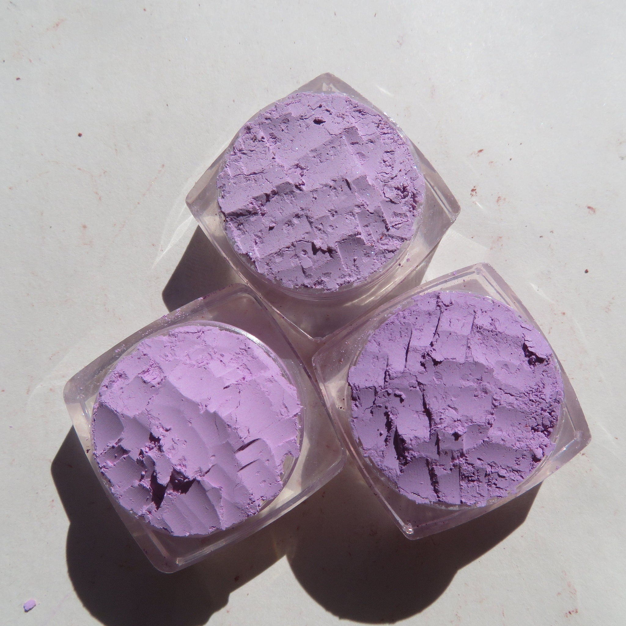Side view of three purple loose mineral eyeshadows in glass jars with black lids, showcasing shimmer texture