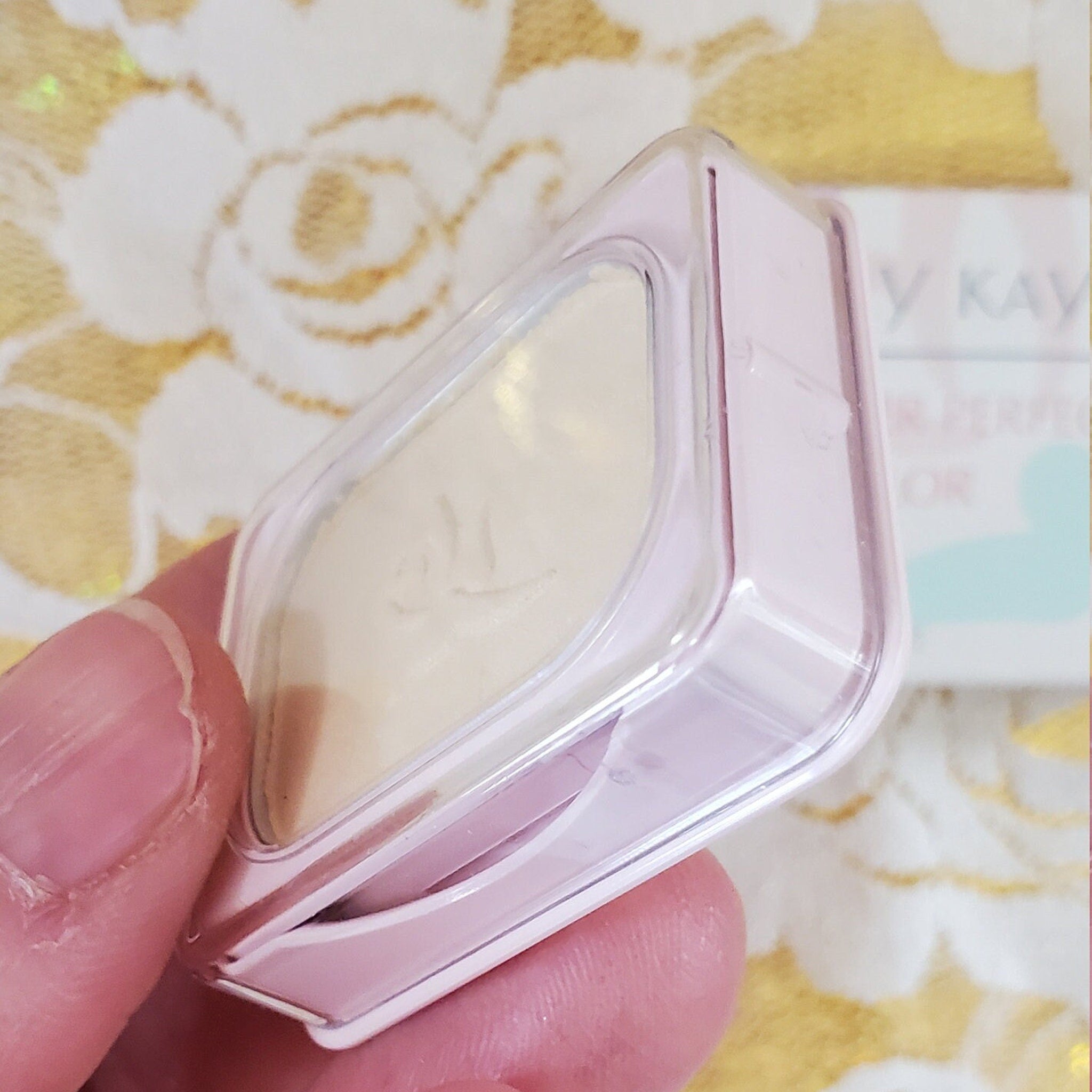 Powder compact in pink and clear plastic, held by a hand, with a floral background.