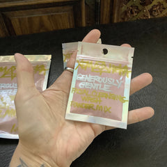 Facial cleansing powder mix in clear iridescent packet, pink powder, held in hand, text visible