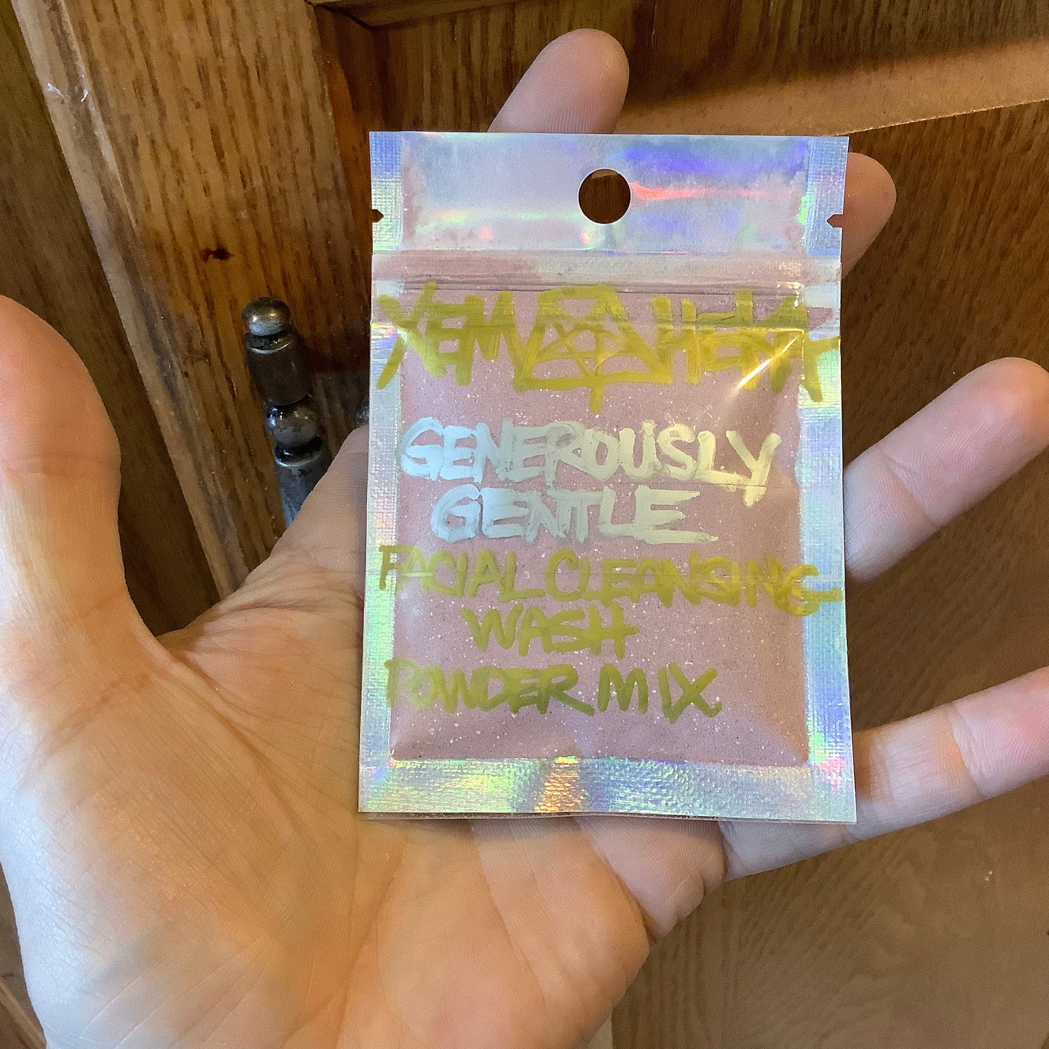 Facial cleansing powder mix in holographic pouch, pink contents, held in hand