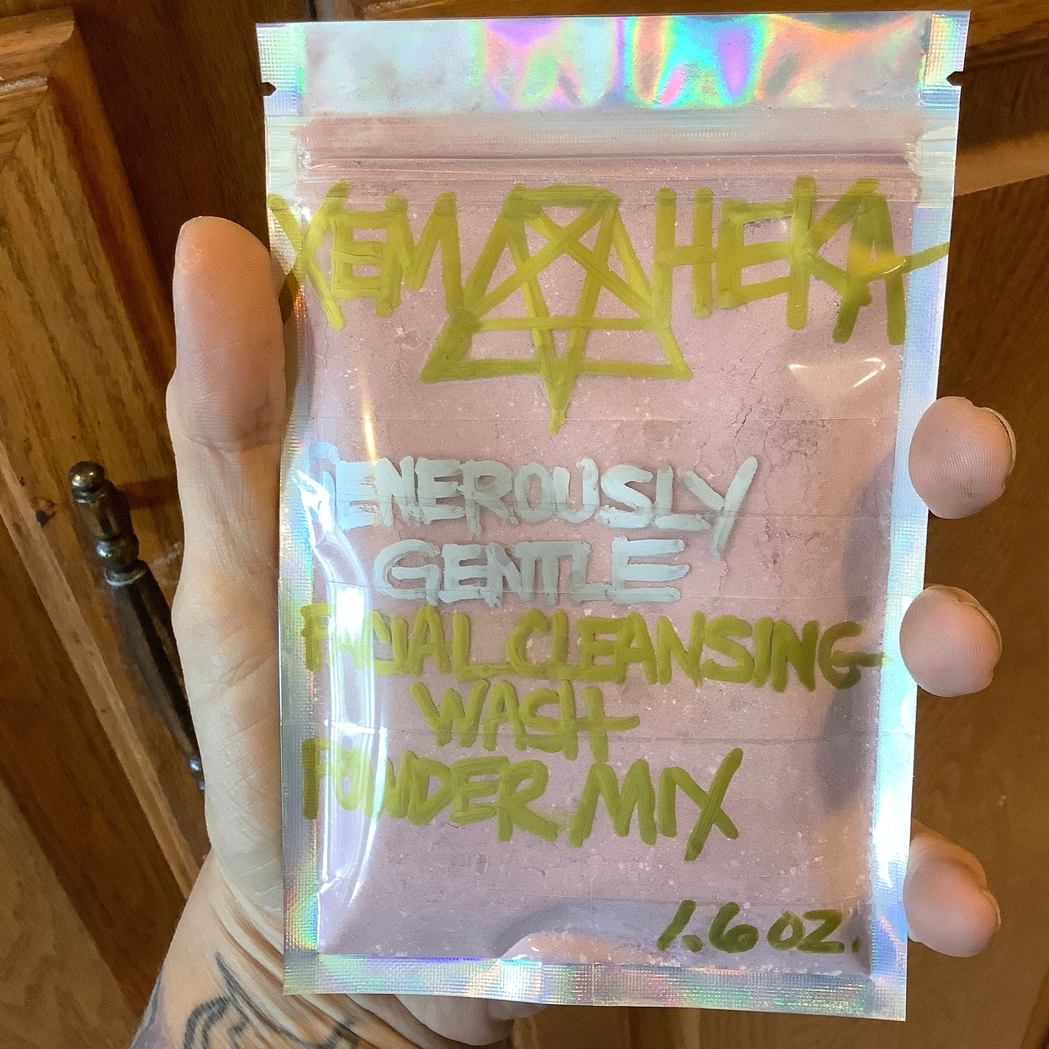 Facial cleansing powder mix in clear iridescent pouch with pink contents and green text, held by hand