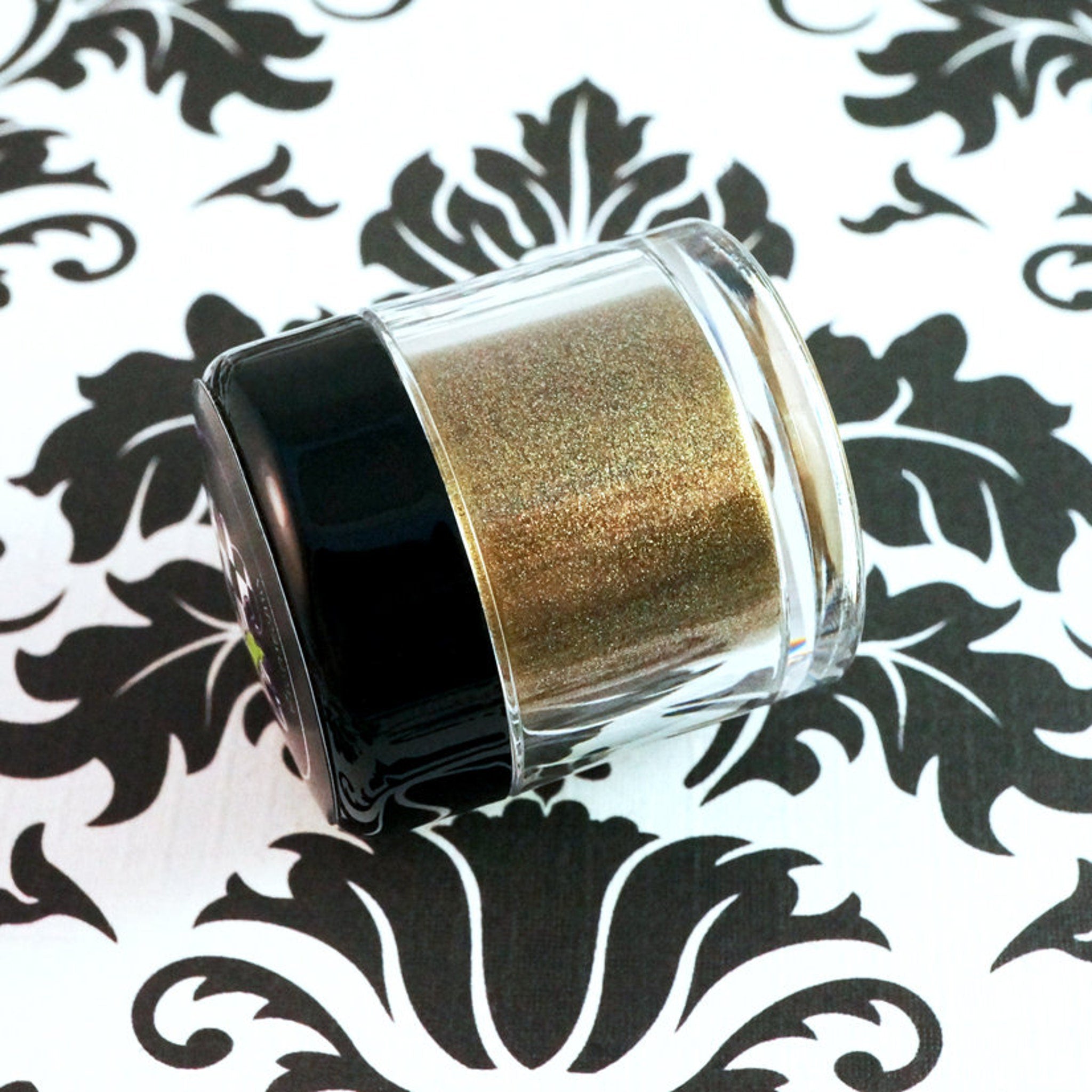 Side view of loose metallic bronze eyeshadow jar showing dark gold powder with green glitter flecks