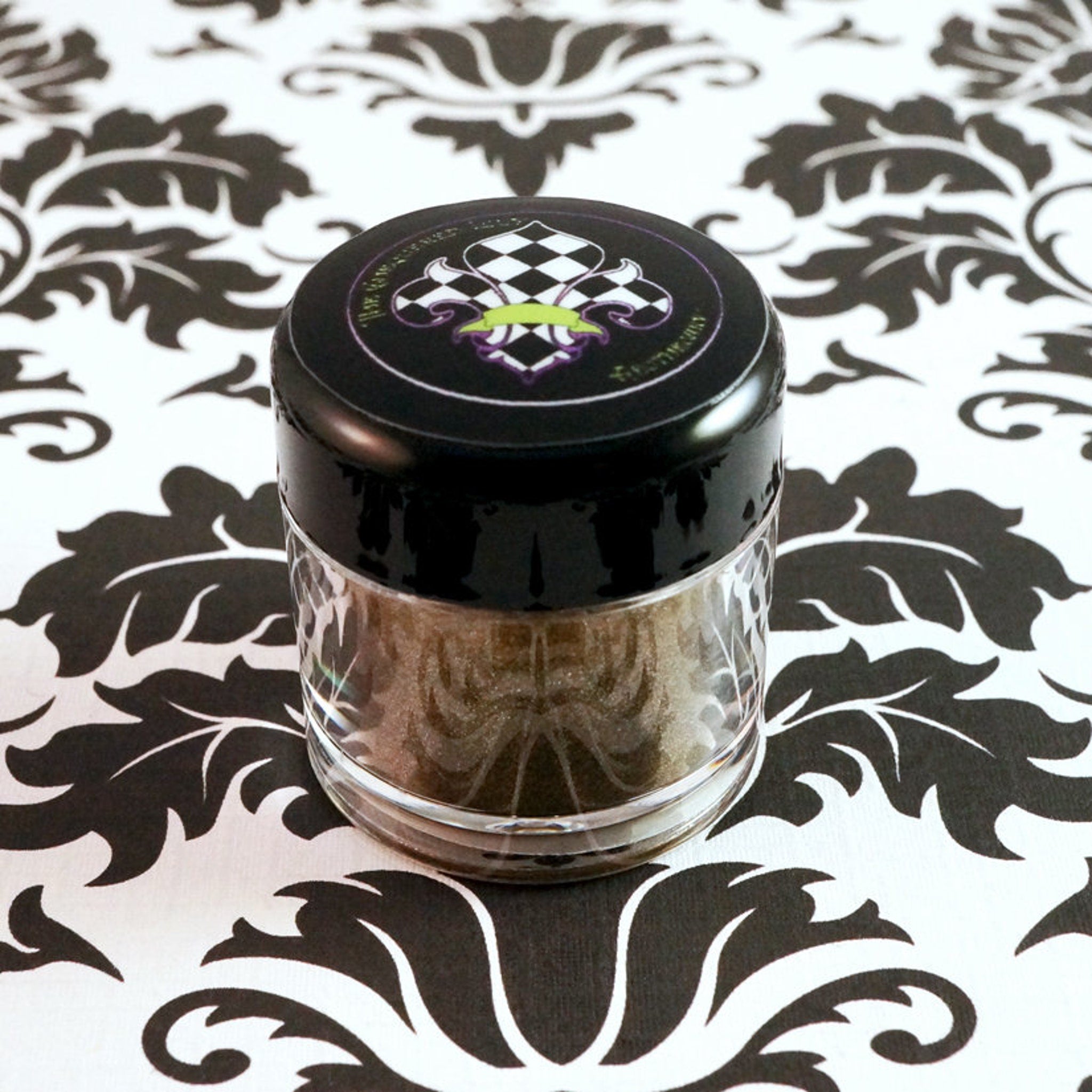 Side view of metallic bronze loose eyeshadow jar showing dark gold powder with green glitter particles