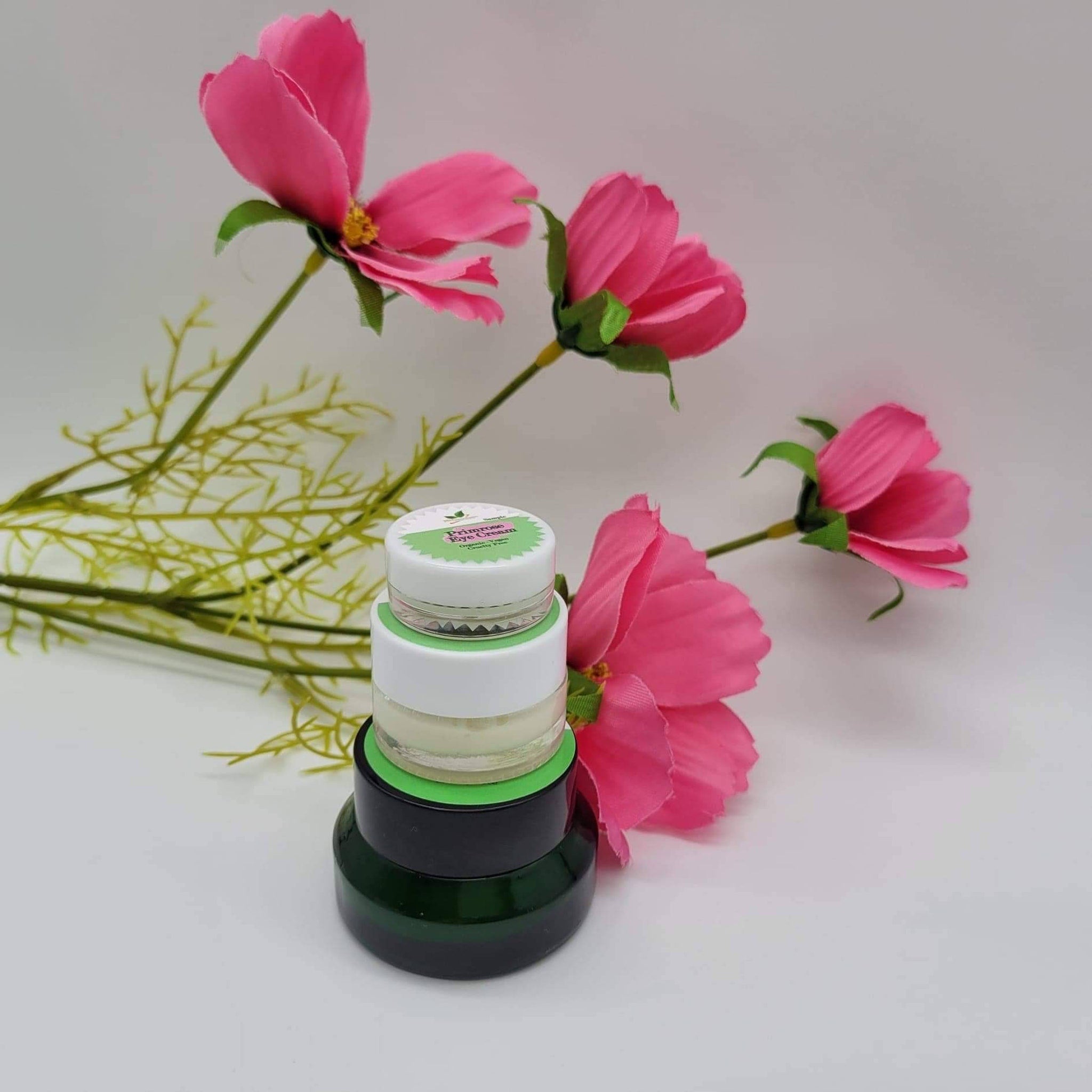 Eye cream jars, stacked green and black glass jars, white lids, and pink flowers in the background