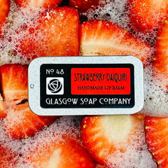 Lip balm tin with red and black label, surrounded by red strawberry slices and bubbles, Glasgow Soap Company