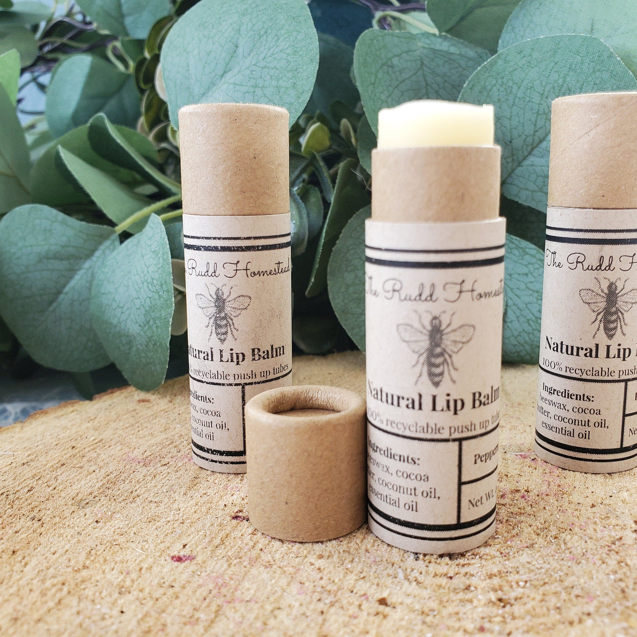 Natural lip balm tubes with brown paper packaging and "The Rudd Homestead" text label on wood surface