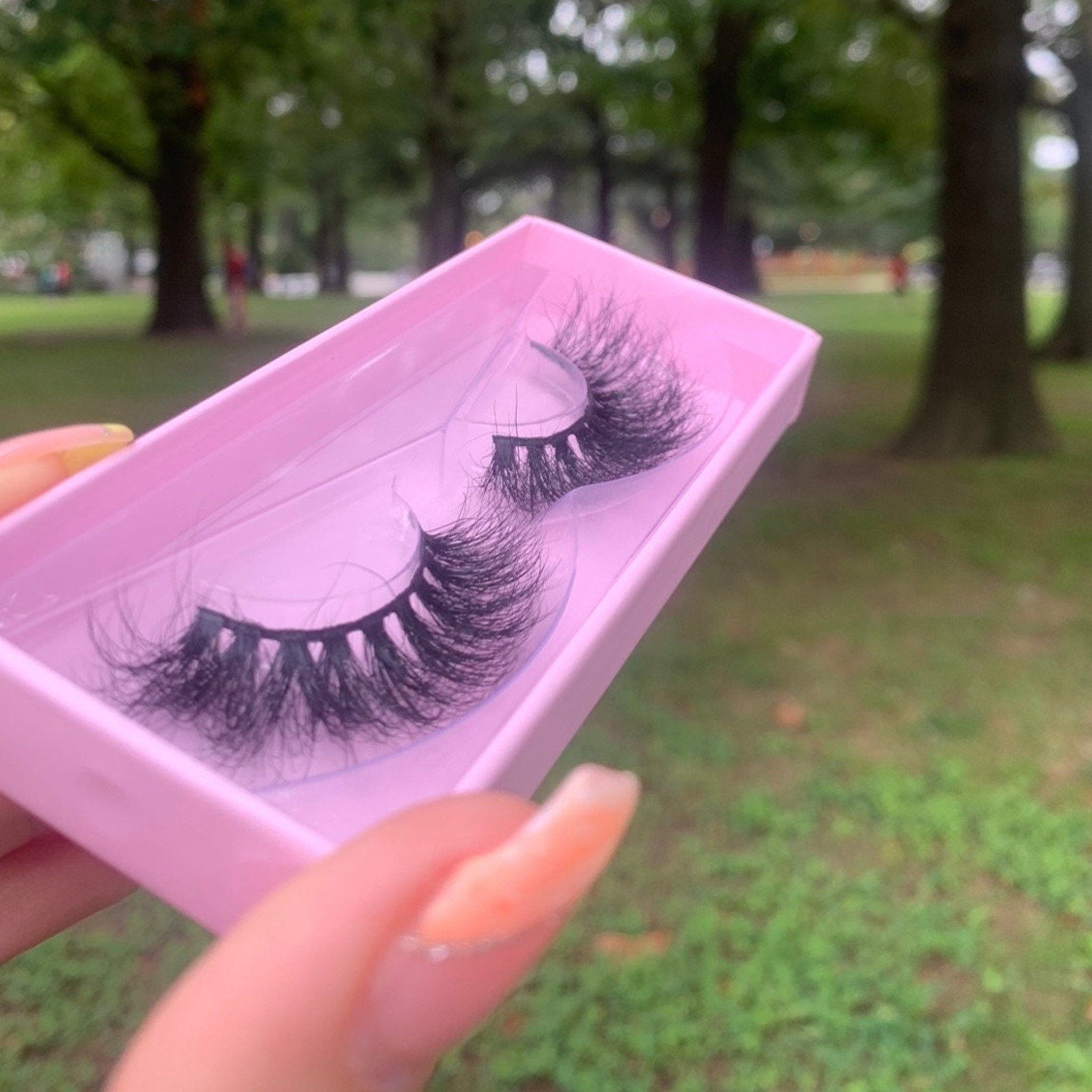 False eyelashes with black synthetic fibers in a pink box held by a hand outdoors