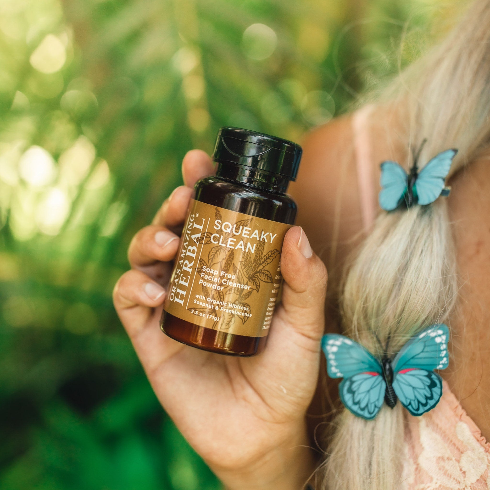 Facial cleanser bottle, brown glass, black cap, held in hand, with Ora's Amazing Herbal label, and butterfly hair clips in background