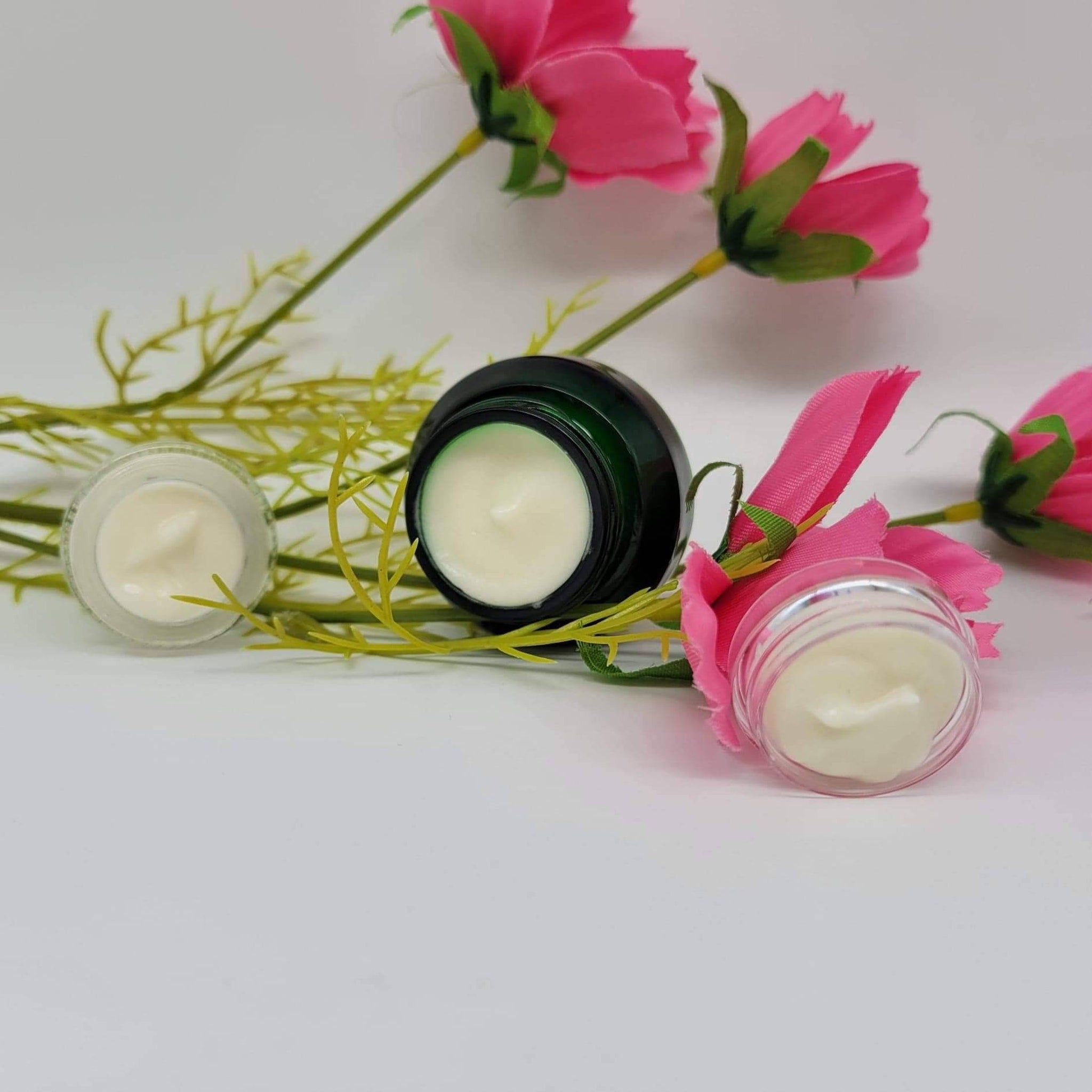 Cream jars with white cream, green and clear jars, pink flowers in background