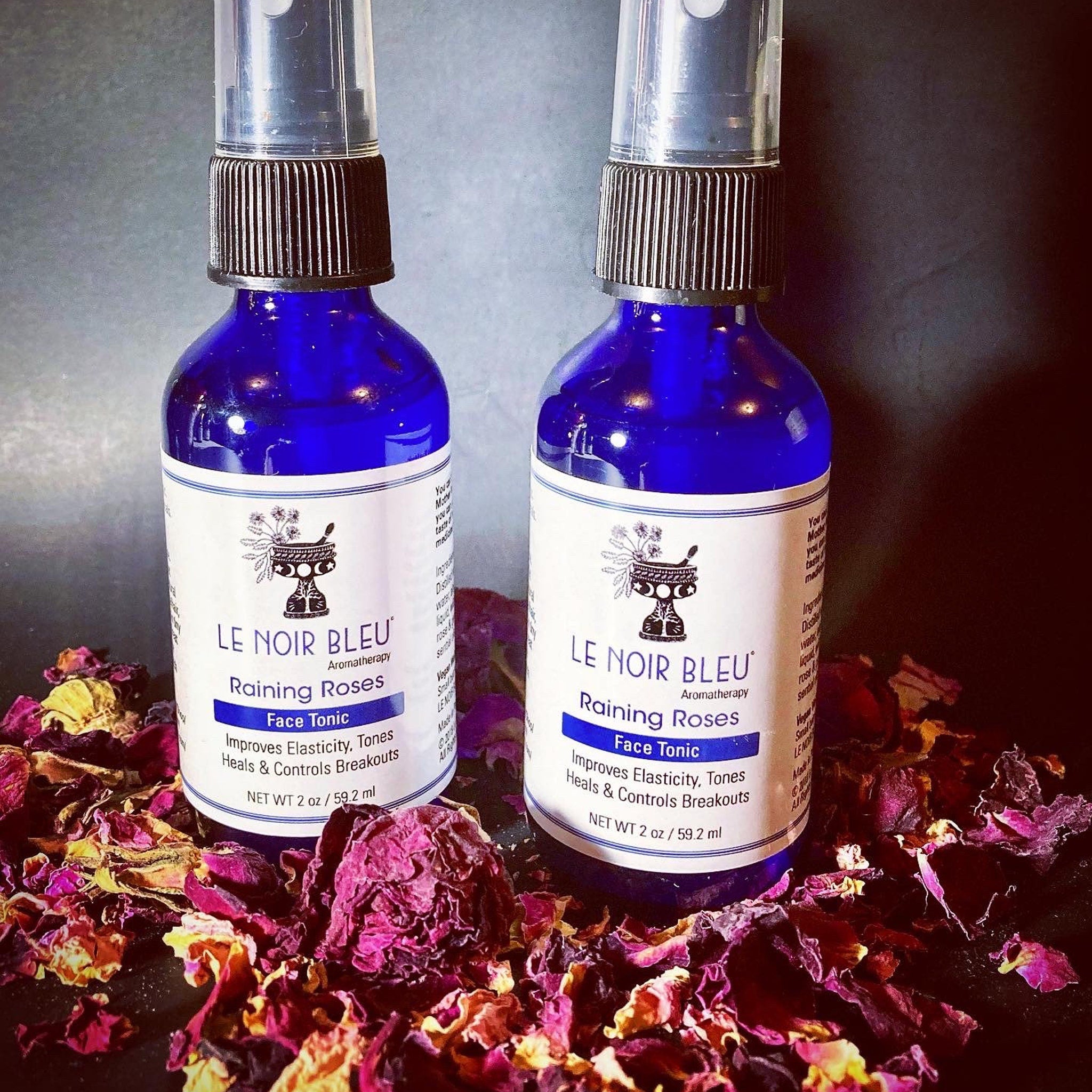 Face tonic bottles, blue glass with black cap, surrounded by rose petals, LE NOIR BLEU branding visible.