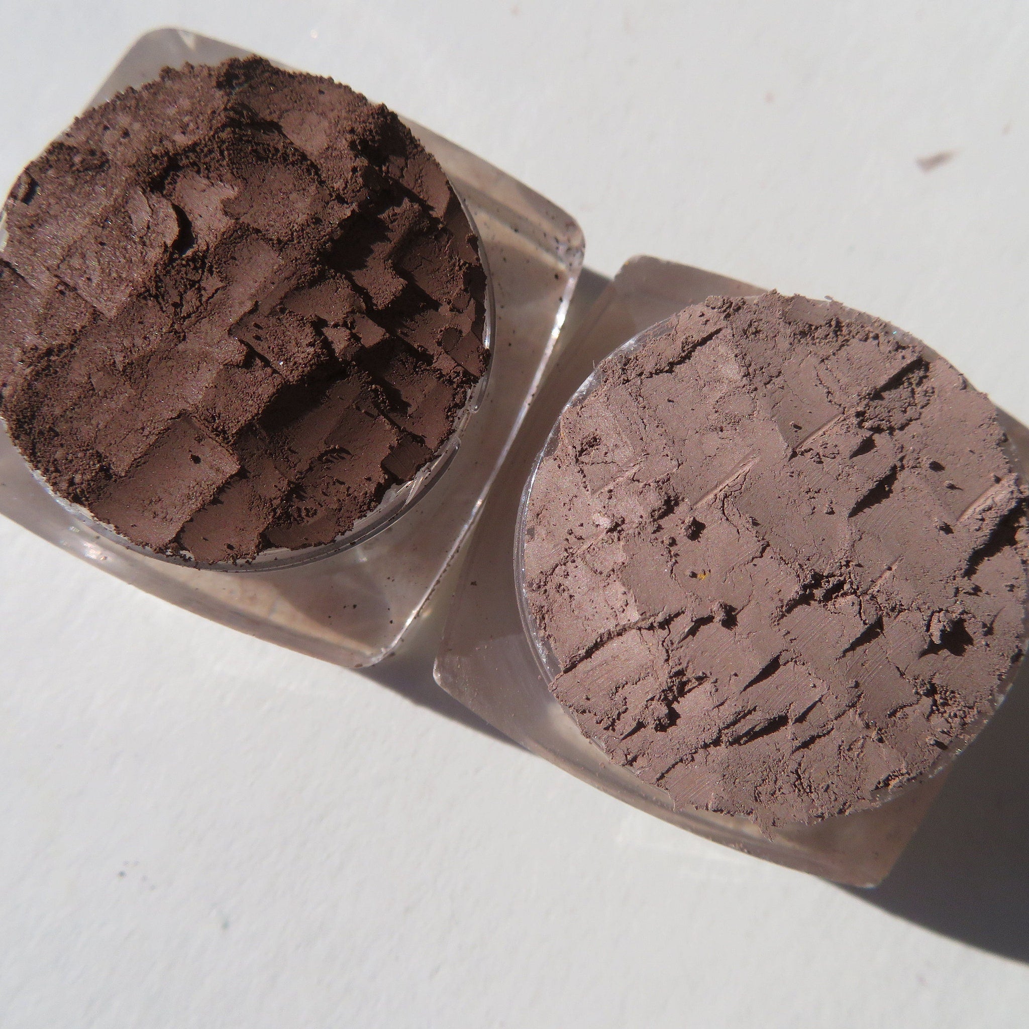Three matte brown loose pigment eyeshadows in sleek black jars with gold lids, arranged on marble surface.