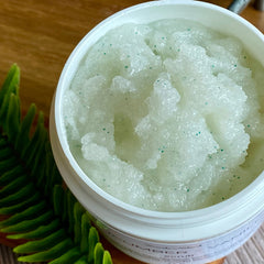 Body scrub with white container, light green granules, and dark green flecks, with fern leaf detail