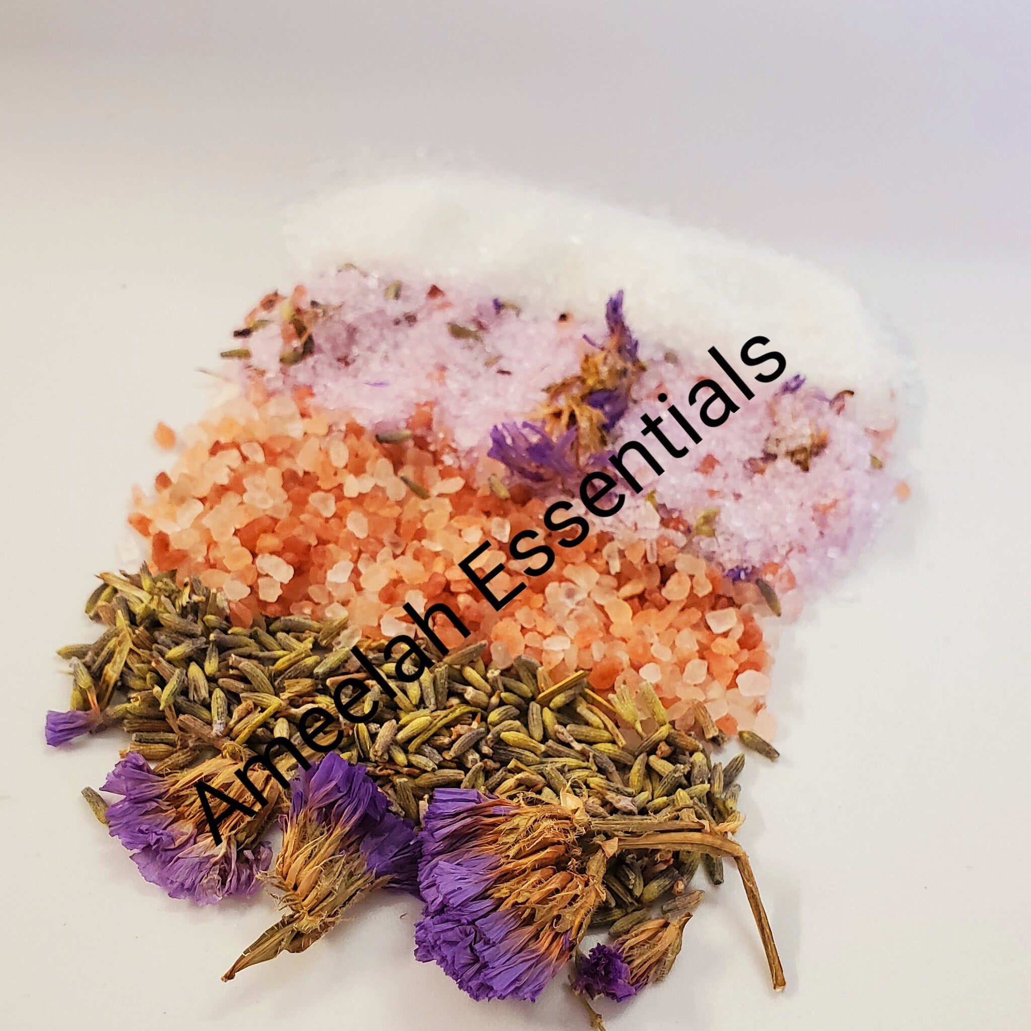 Bath salts with white, pink, and orange granules, dried purple flowers, and Ameelah Essentials text