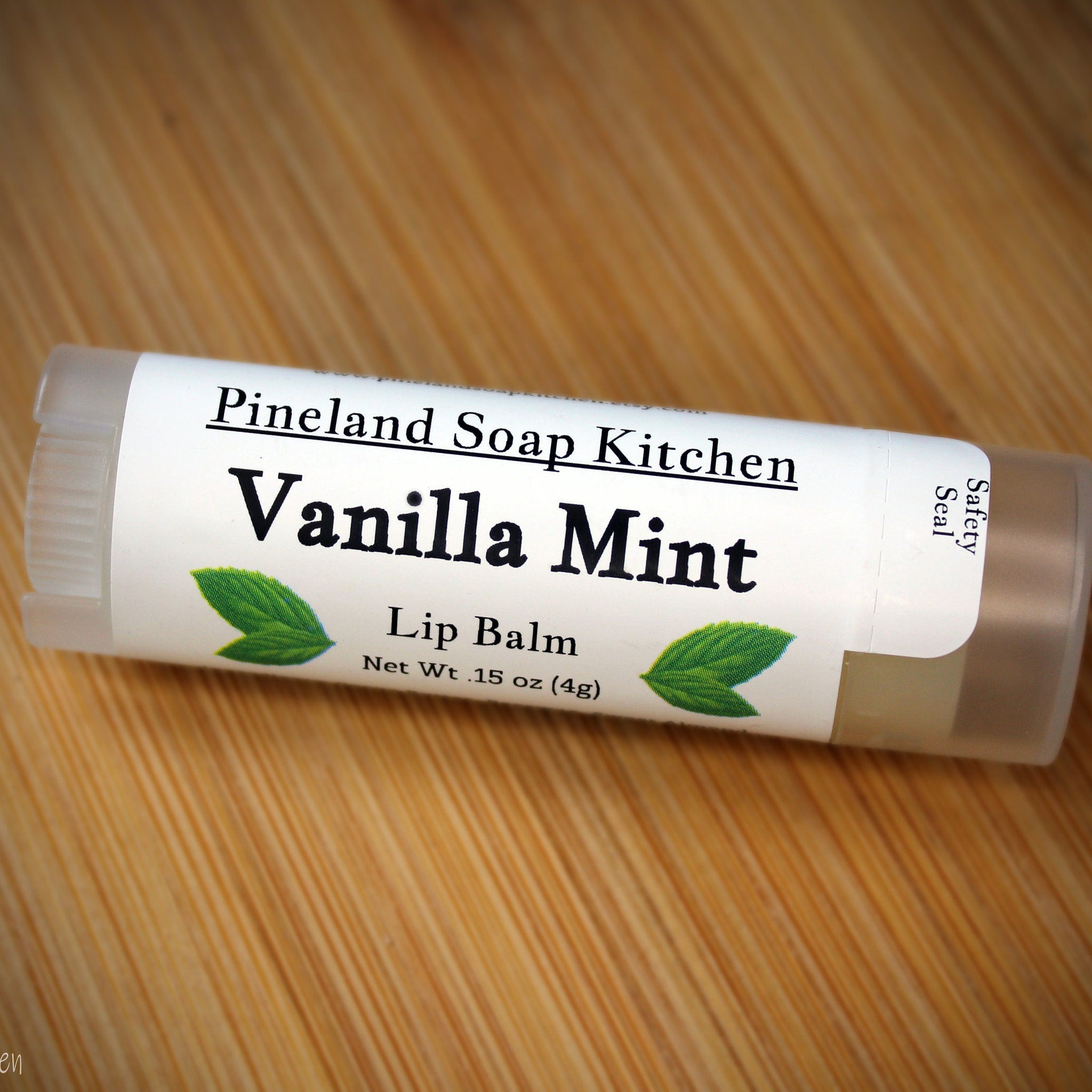 Lip balm tube with white label, green leaf design, and "Vanilla Mint" text on wooden surface
