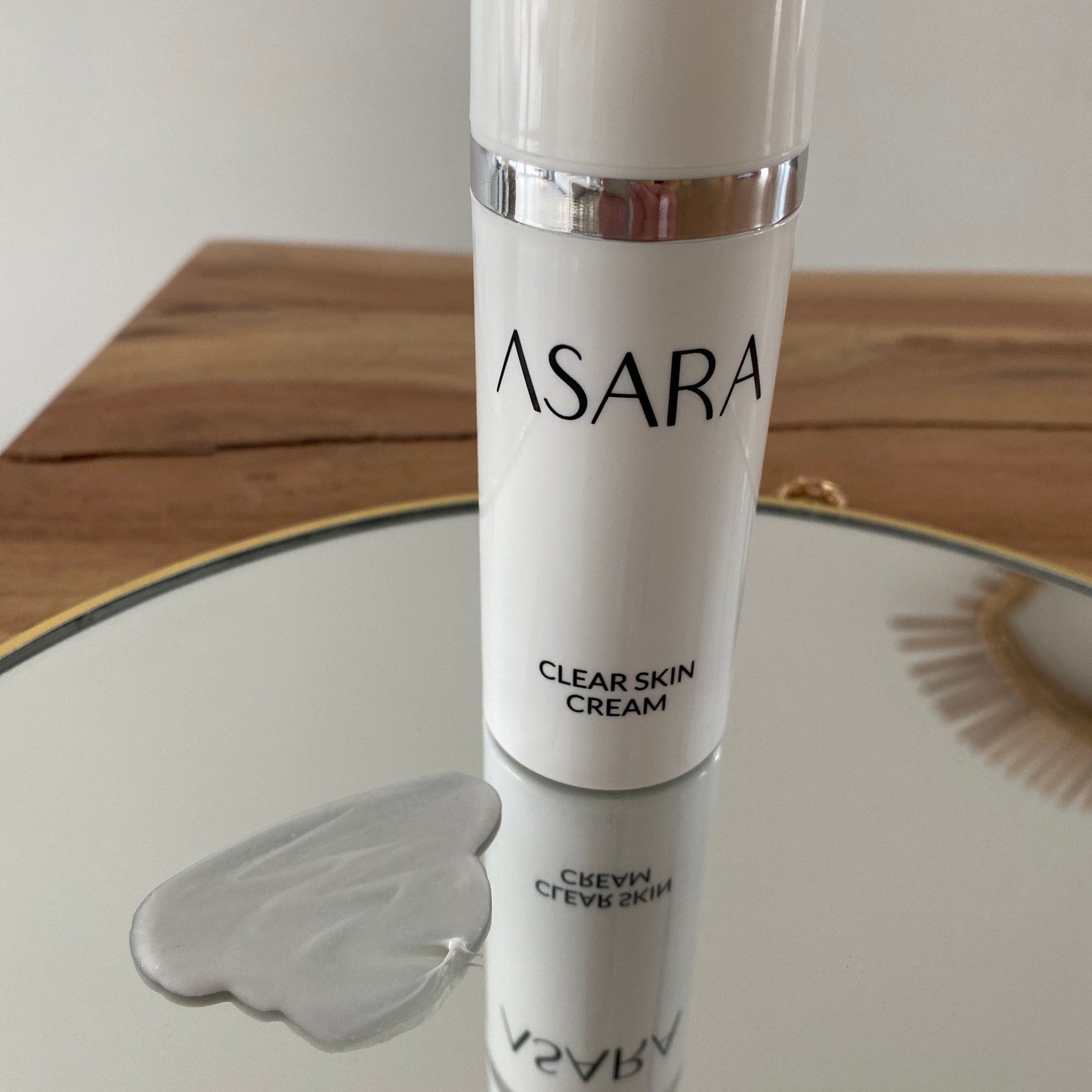 Clear skin cream bottle, white with silver band, ASARA brand visible on bottle, reflected on mirror surface