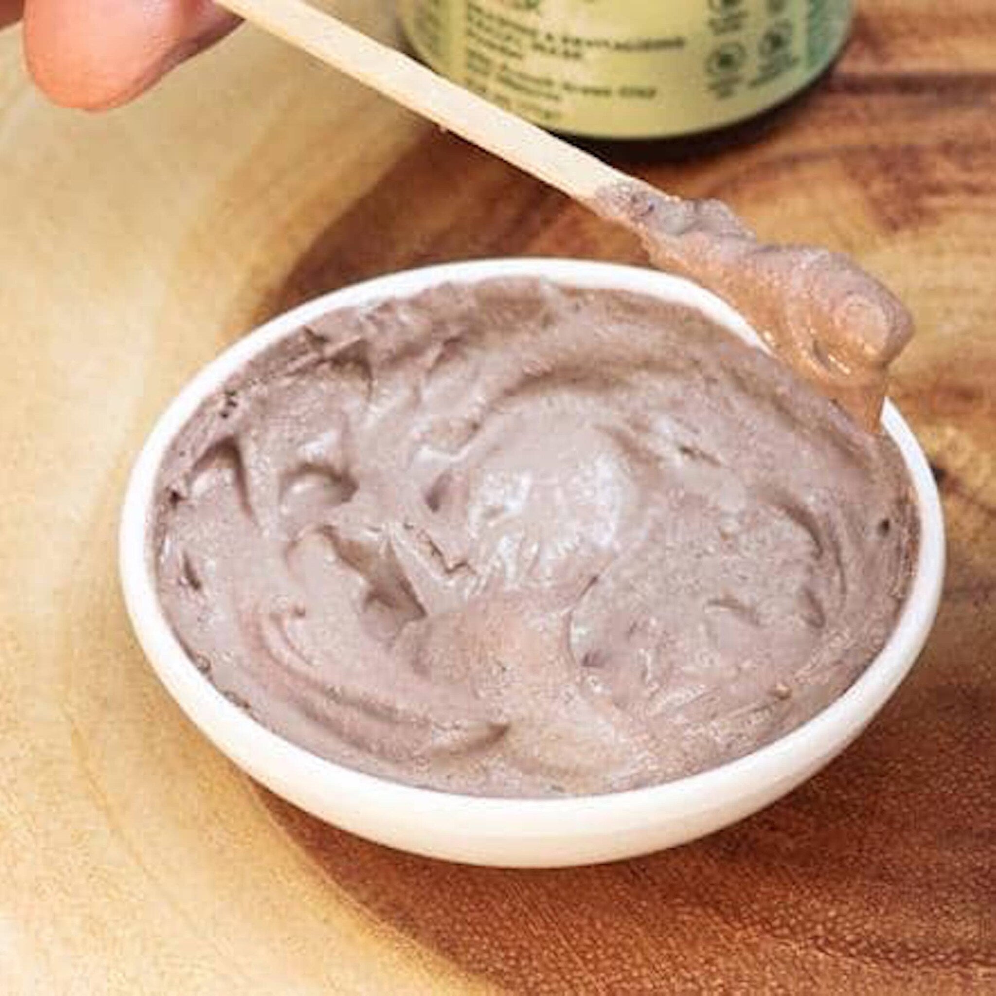Face mask in a white bowl with brown cream and wooden applicator on a wooden surface