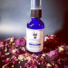 Toner bottle on black surface, blue glass bottle with black cap, surrounded by rose petals, Le Noir Bleu label visible
